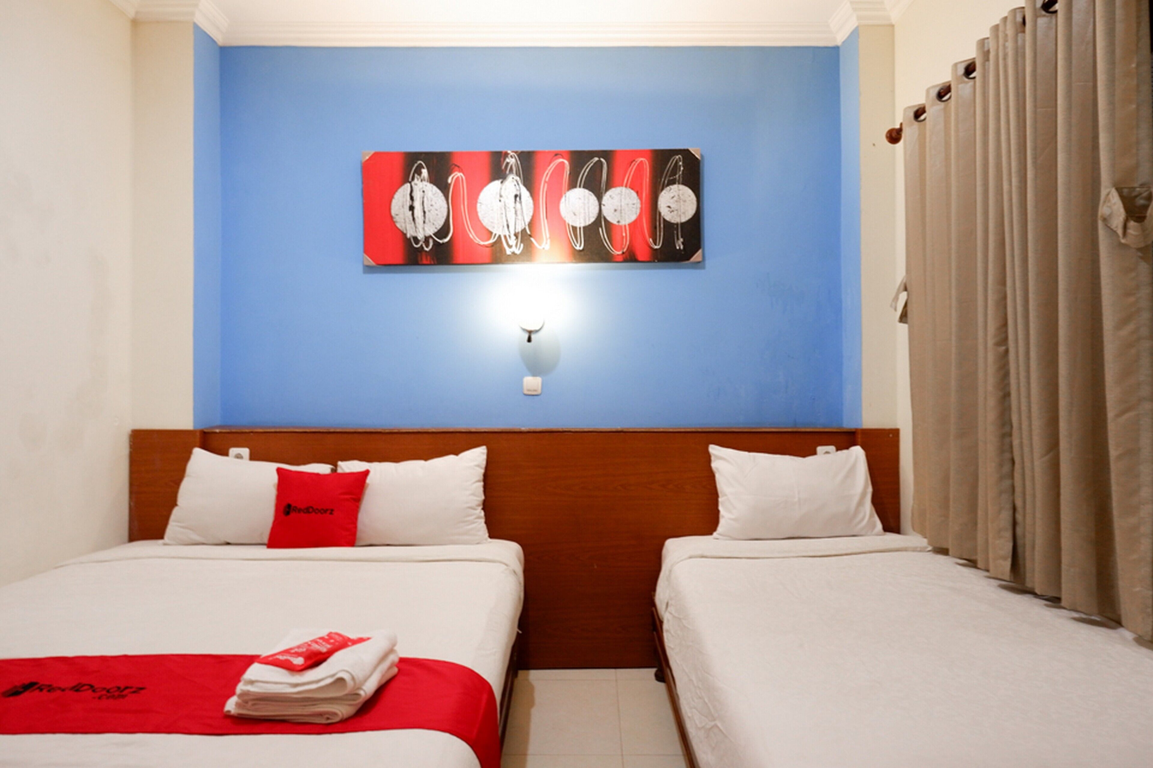 A hotel room with two beds, a blue accent wall, a modern artwork, and a red blanket on one bed.