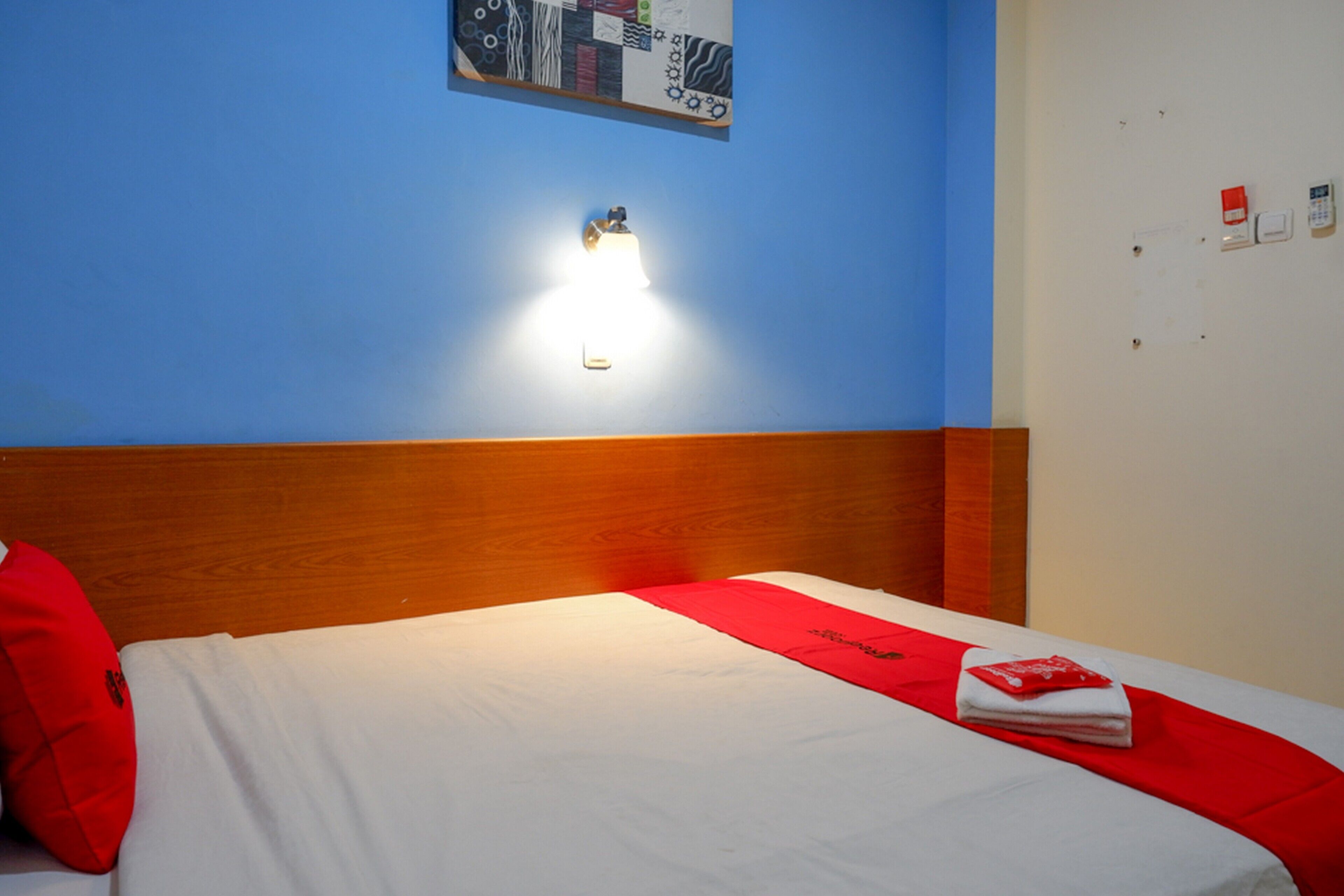 A hotel room with a wooden headboard, a bed with white bedding and a red accent, a wall-mounted light, and a bulletin board with various items pinned to it.