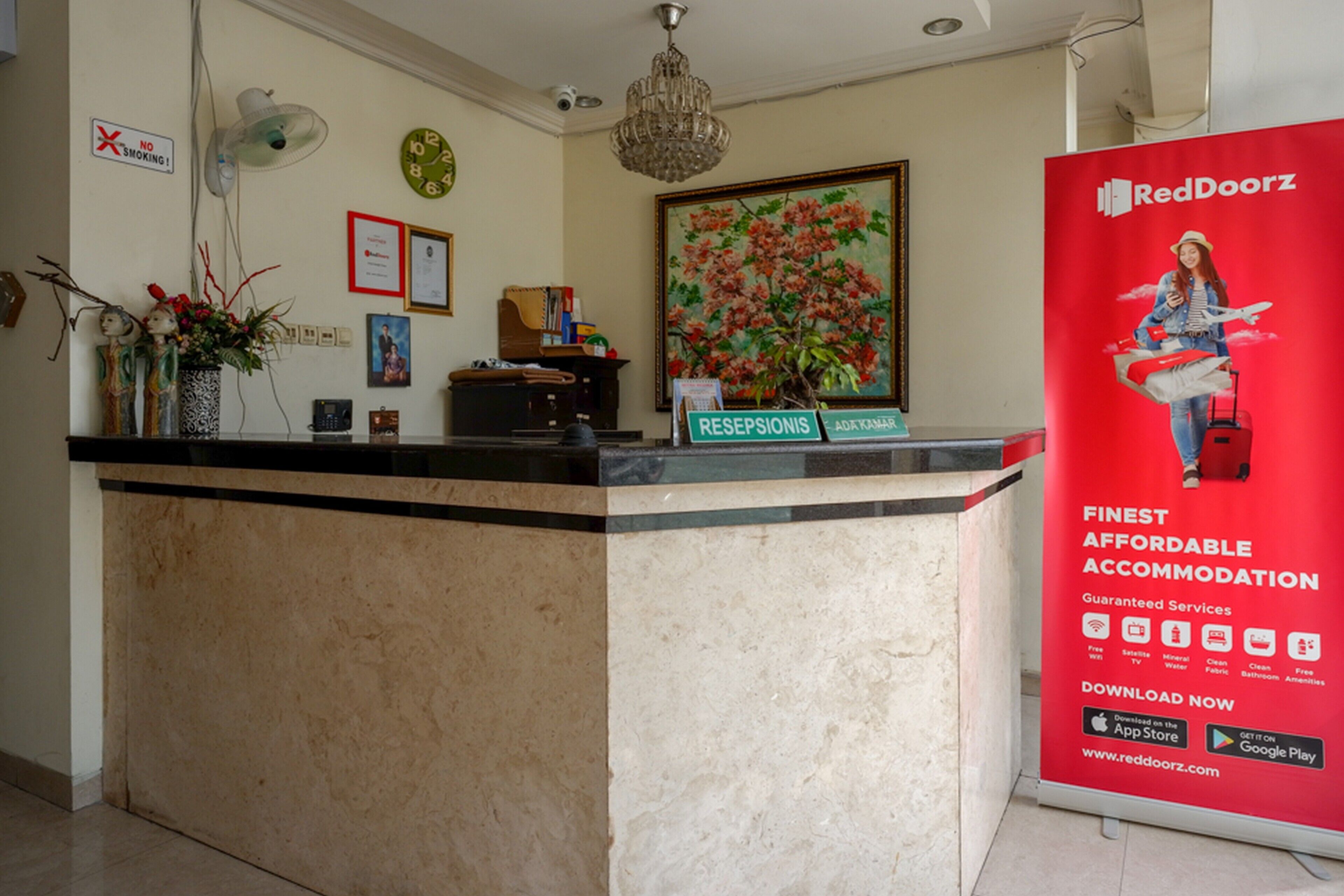 A reception desk with a 'No Smoking' sign, a clock, framed certificates, a chandelier, and a RedDoorz advertisement.