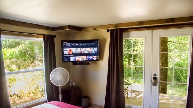 Ein Hotelzimmer mit einem Bett, einem an der Wand montierten Fernseher, einem Ventilator und großen Fenstern mit Vorhängen.