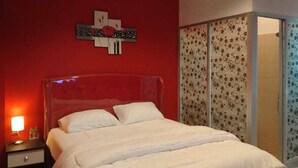 A bedroom with a red wall, a bed with white bedding, a nightstand with a lamp, and a sliding door with patterned curtains. - Reddoorz @ Sindangbarang Bogor (Bogor)