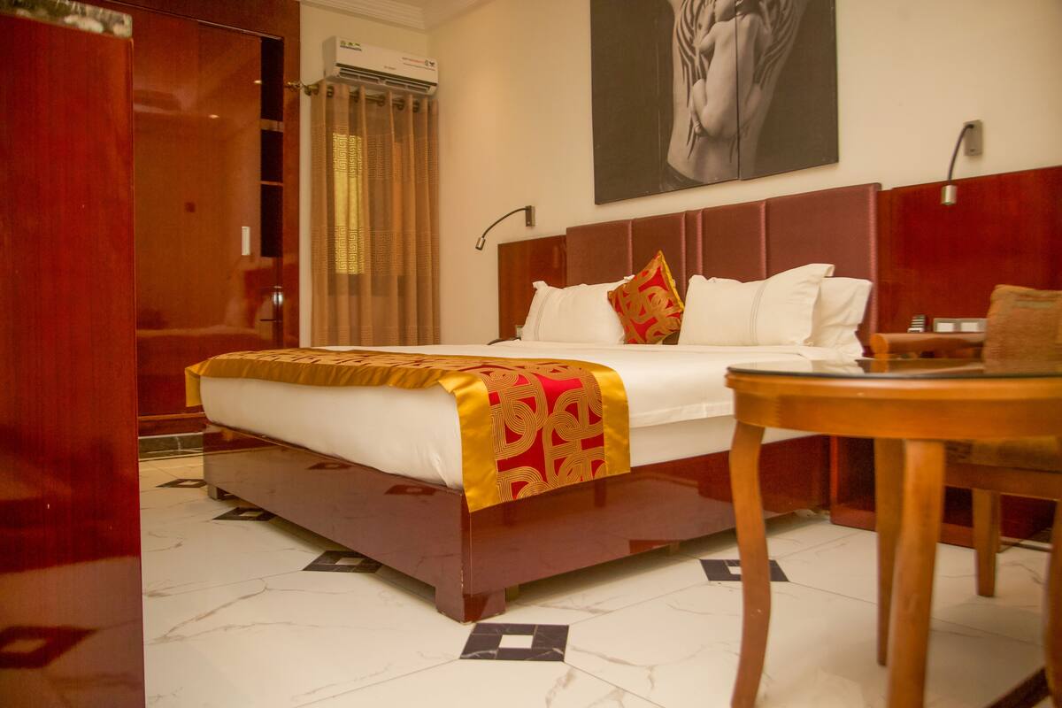 A hotel room with a large bed, a wooden headboard, a bedside table, and a wall-mounted air conditioner.