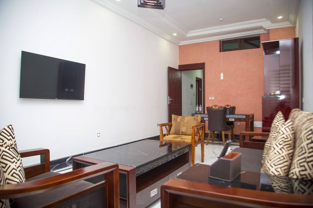 A modern living room with a large flat-screen TV mounted on the wall, a glass-top coffee table, and wooden furniture.