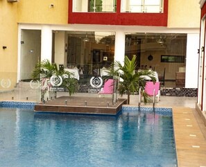 A swimming pool with a wooden deck, palm trees, and a building with a red and white facade in the background. - Hotel Capitol Abidjan (Abidjan)