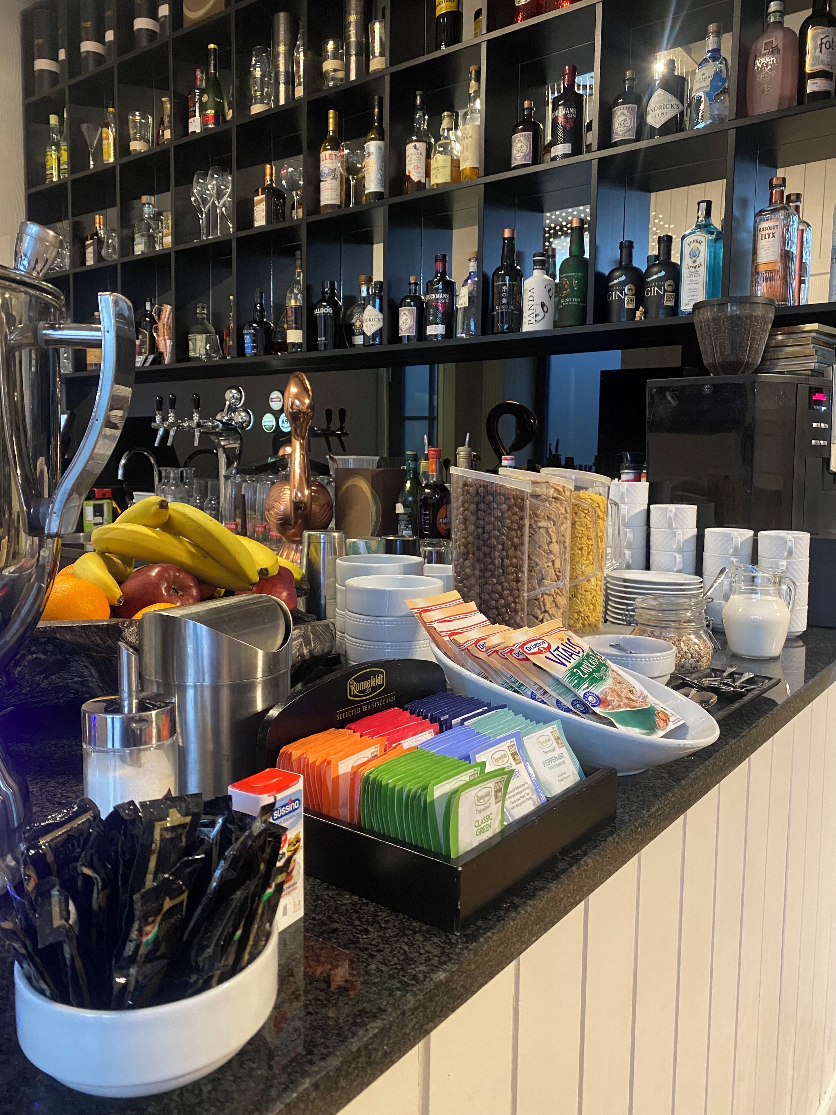 A bar area with shelves of liquor bottles, a coffee machine, and various food items like bananas and cereal.
