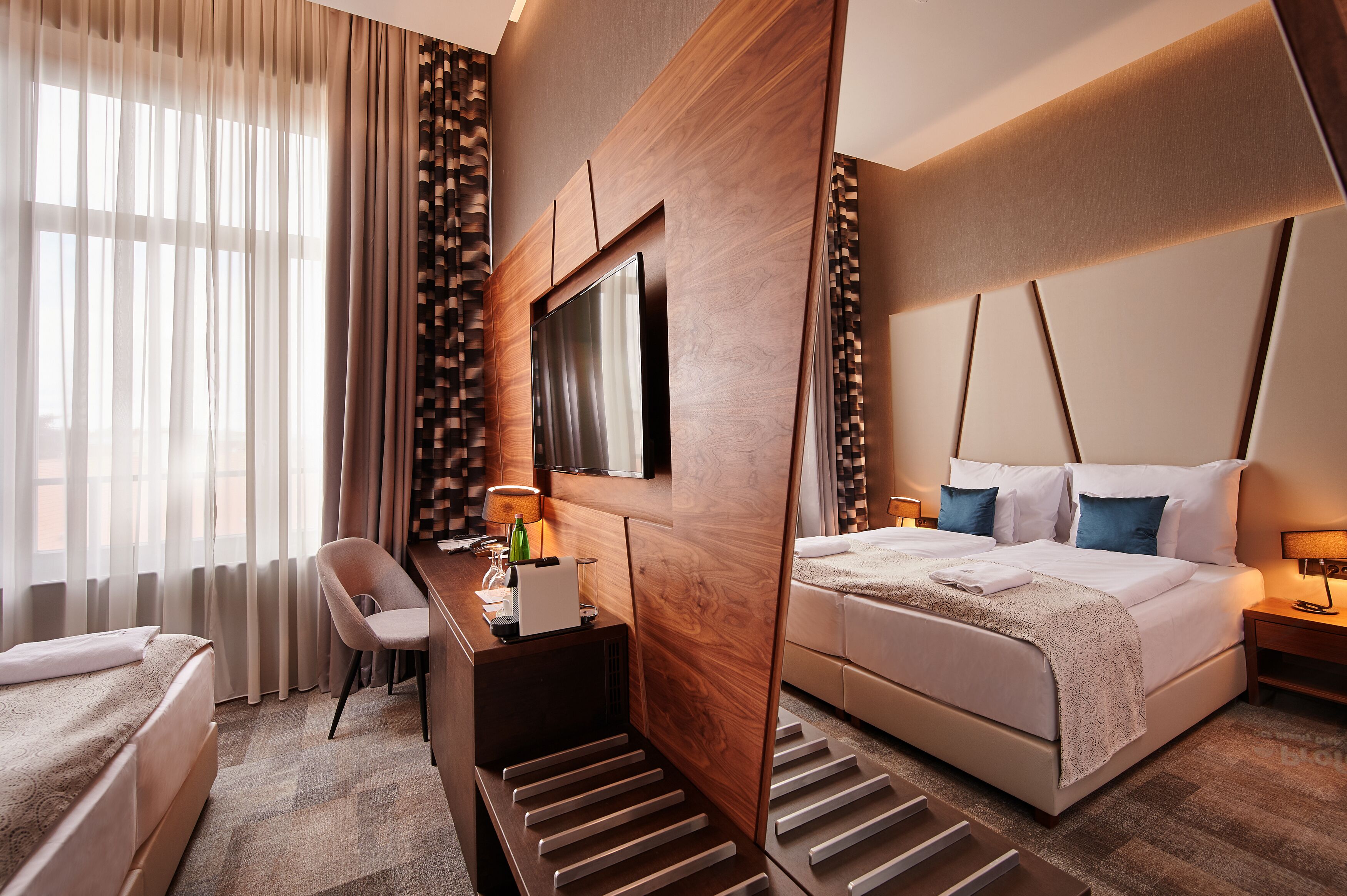 A modern hotel room with a large bed, a desk with a chair, a television, and a window with sheer curtains.