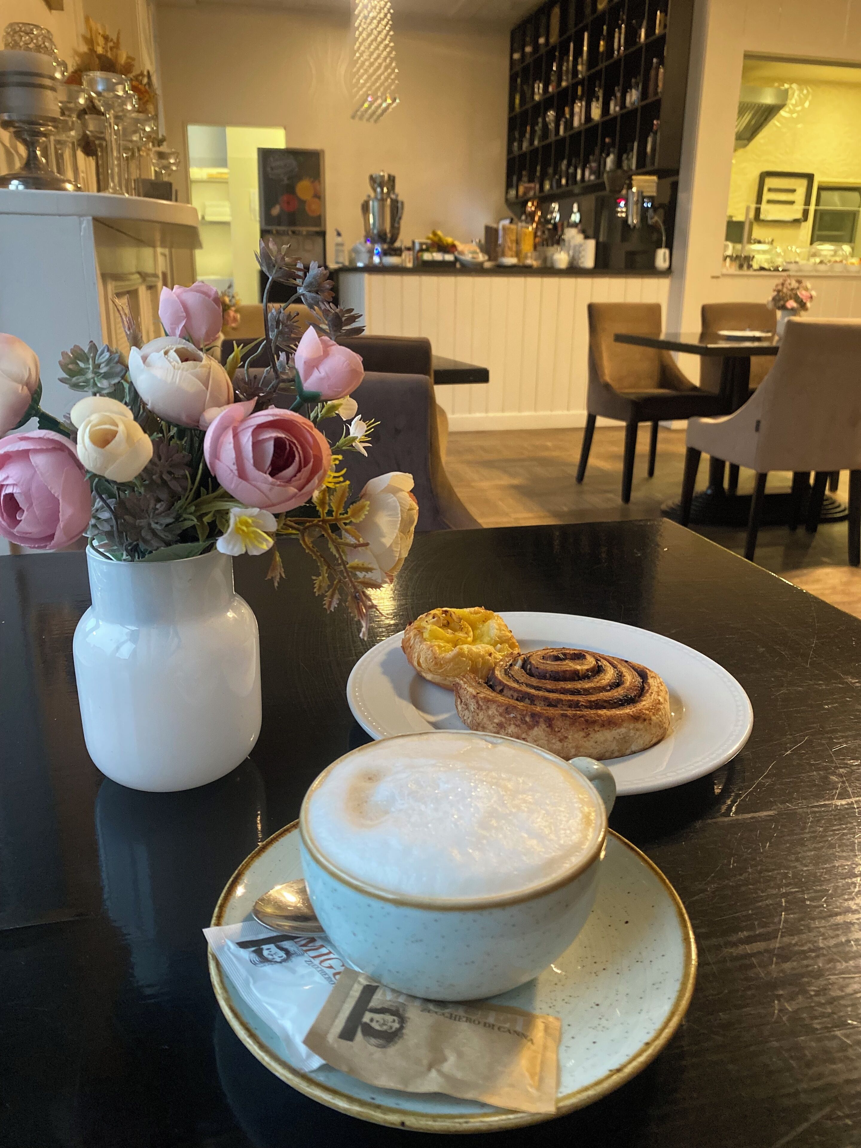 Ein Esstisch mit einer weißen Vase voller rosa und weißer Blumen, einem Teller mit einem Zimtschnecke und einer Kaffeetasse mit Sahnezucker.