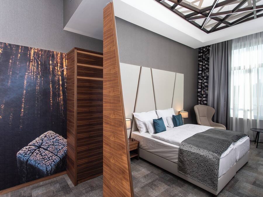 Ein modernes Hotelzimmer mit einem großen Bett, einem Holzschrank, einem Nachttisch, einem Sessel und einem kleinen Tisch.