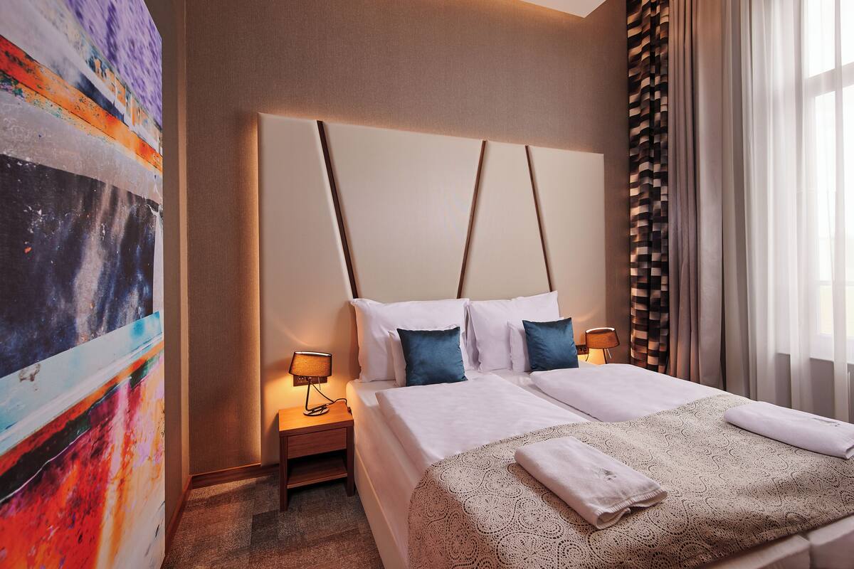 Una habitación de hotel con una cama grande, una mesita de noche con lámpara, un cuadro abstracto colorido en la pared y una ventana con cortinas.
