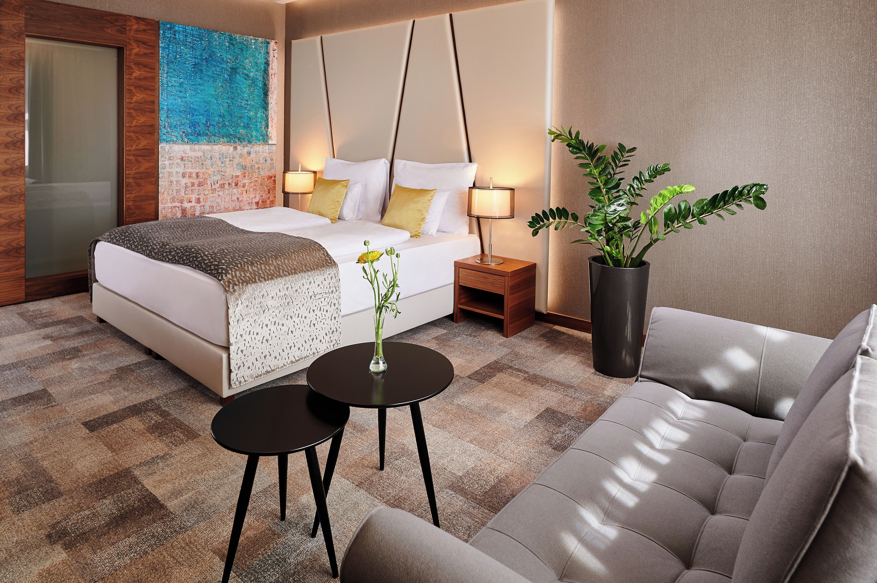 A modern hotel room with a bed, bedside tables, a sofa, and a potted plant.