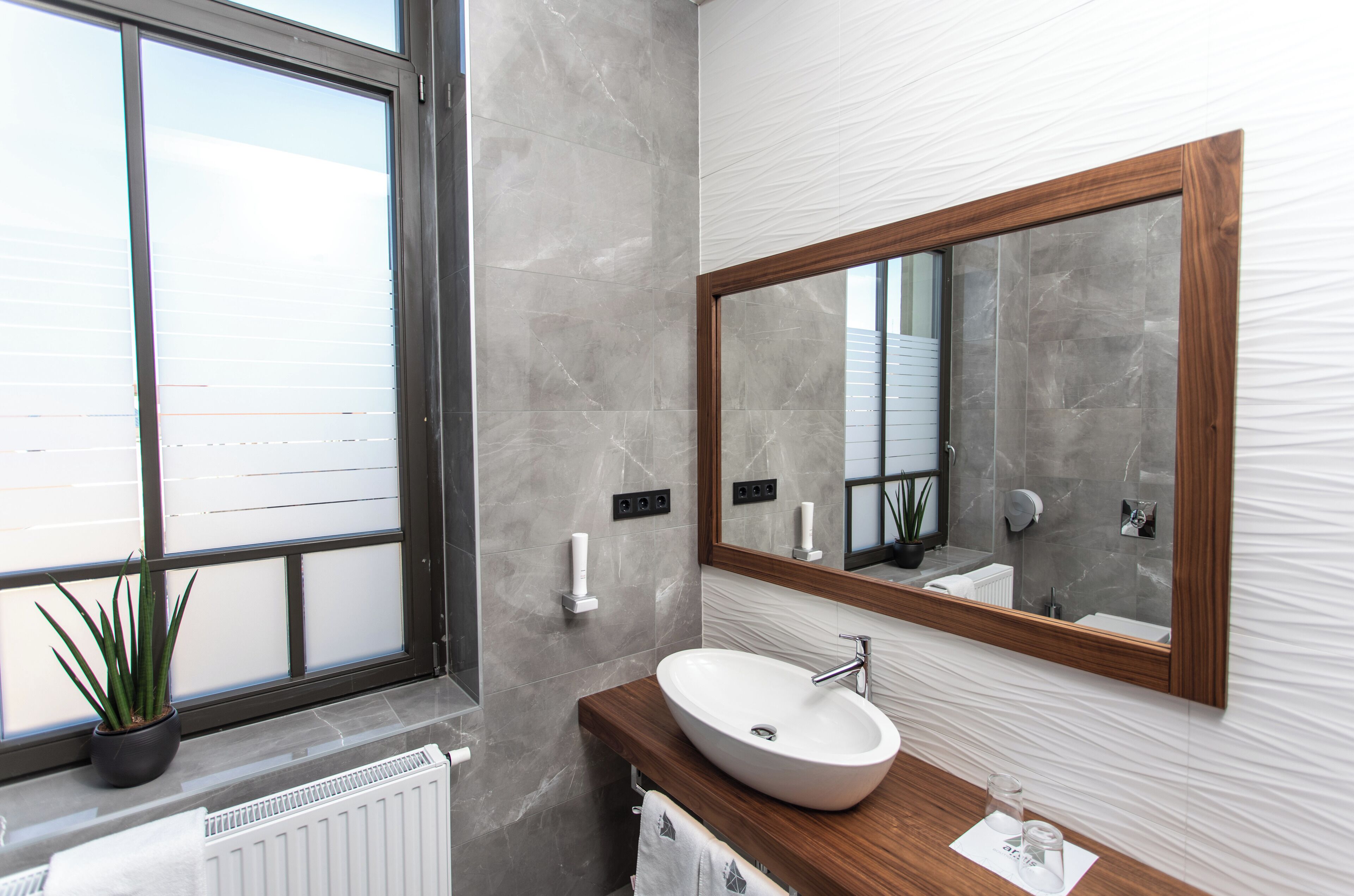 Un baño moderno con un ventanal grande, un tocador de madera, un lavabo blanco y un espejo con marco de madera.