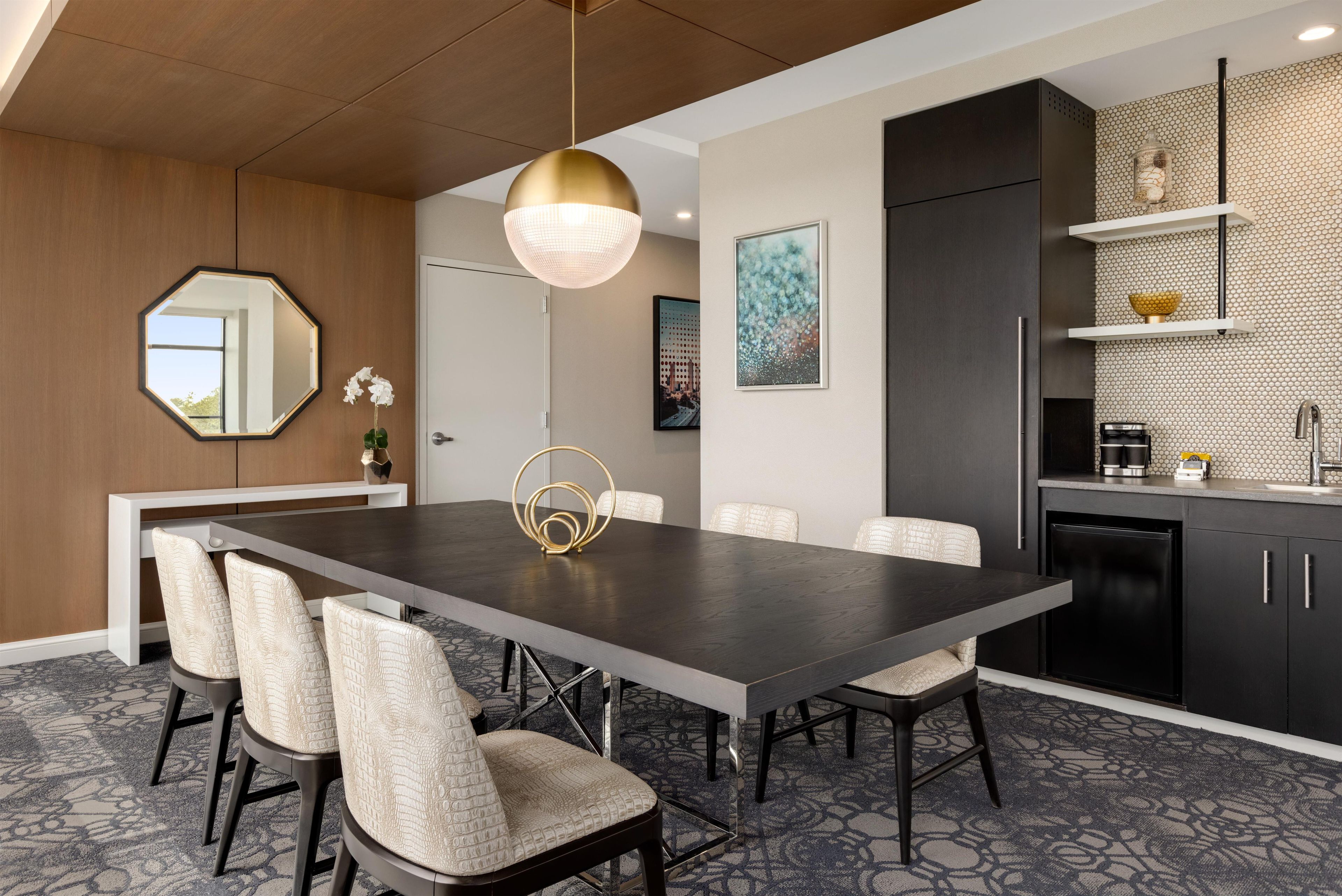 A modern dining area with a dark wooden table, six chairs, a pendant light, and a kitchen with dark cabinets and a tiled backsplash.