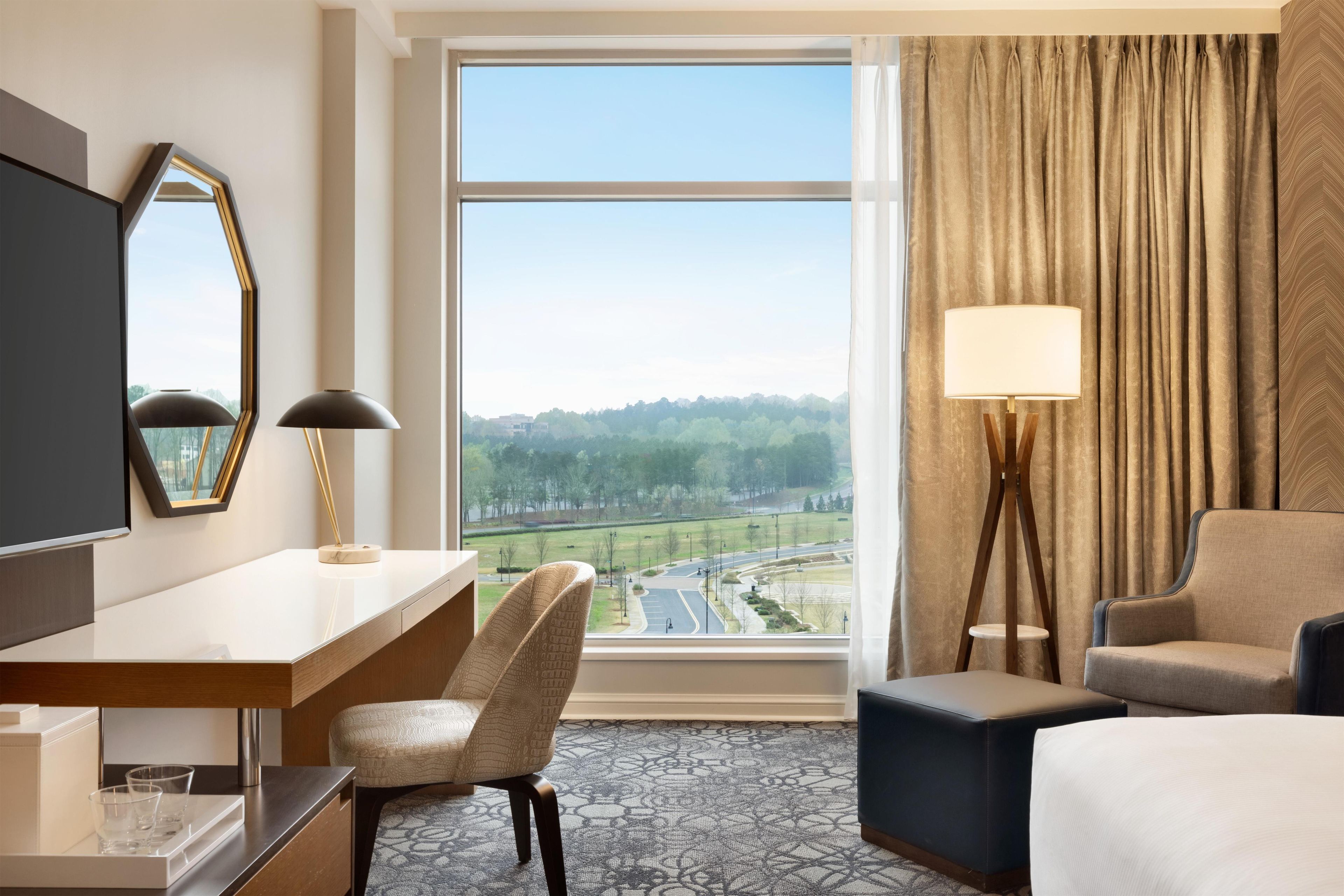 Habitación de hotel con escritorio, silla, cama, televisor, ventana con vista a un paisaje y una lámpara.