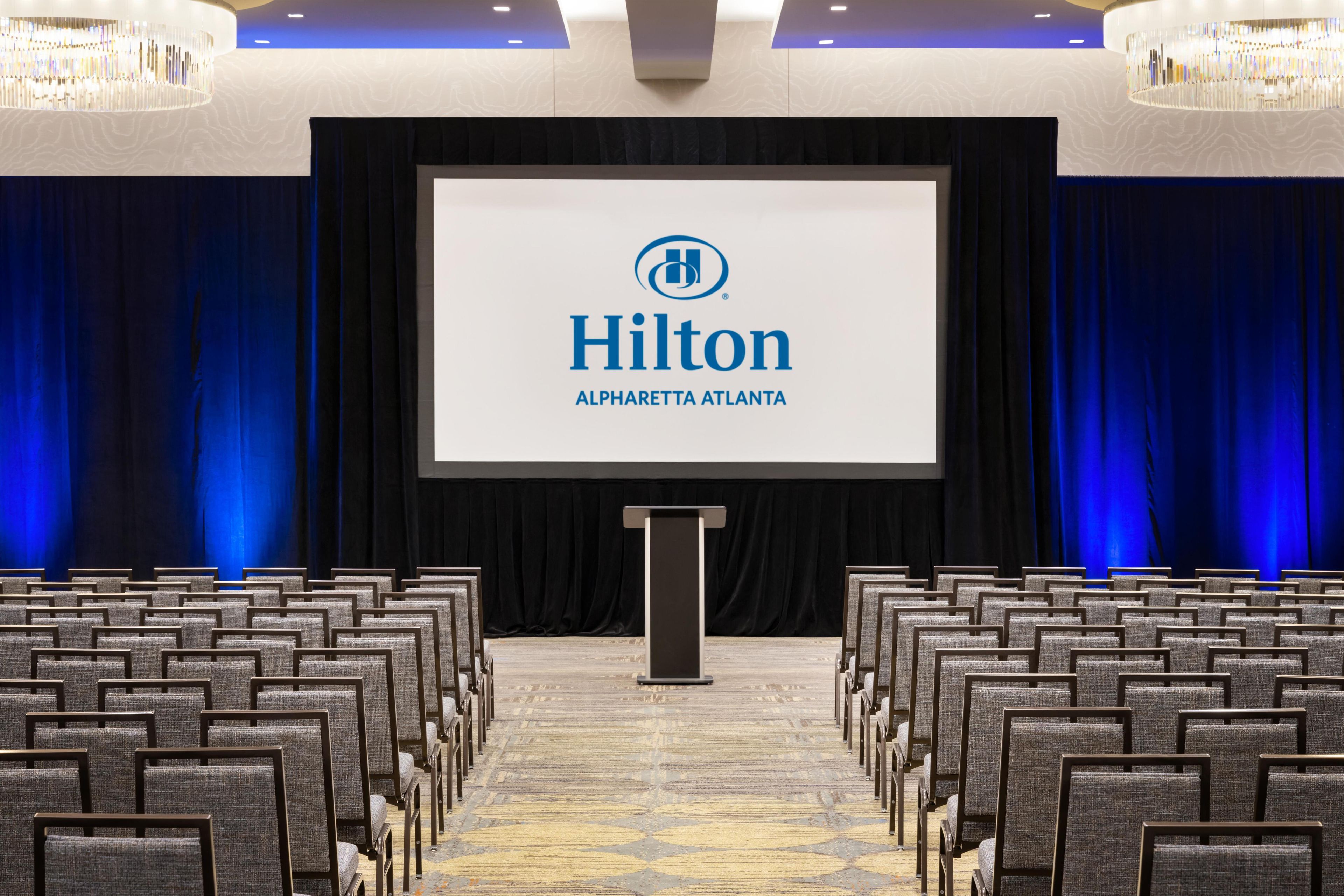 A conference room with rows of chairs, a podium, and a large screen displaying the Hilton logo.