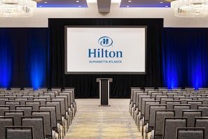 A conference room with rows of chairs, a podium, and a large screen displaying the Hilton logo. - Hilton Alpharetta Atlanta (Alpharetta)
