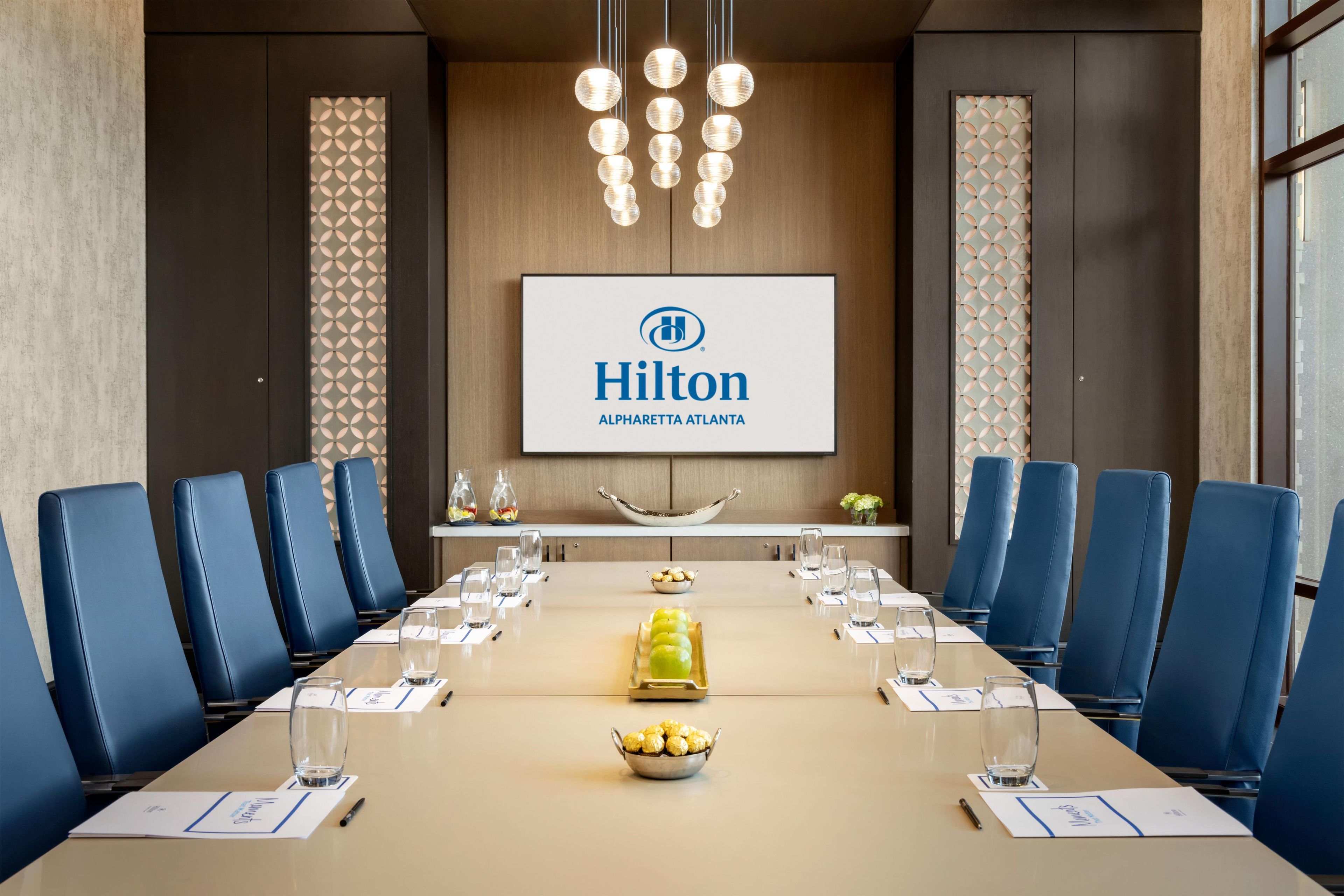 A conference room with a long table, blue chairs, a Hilton logo, and a decorative wall panel.