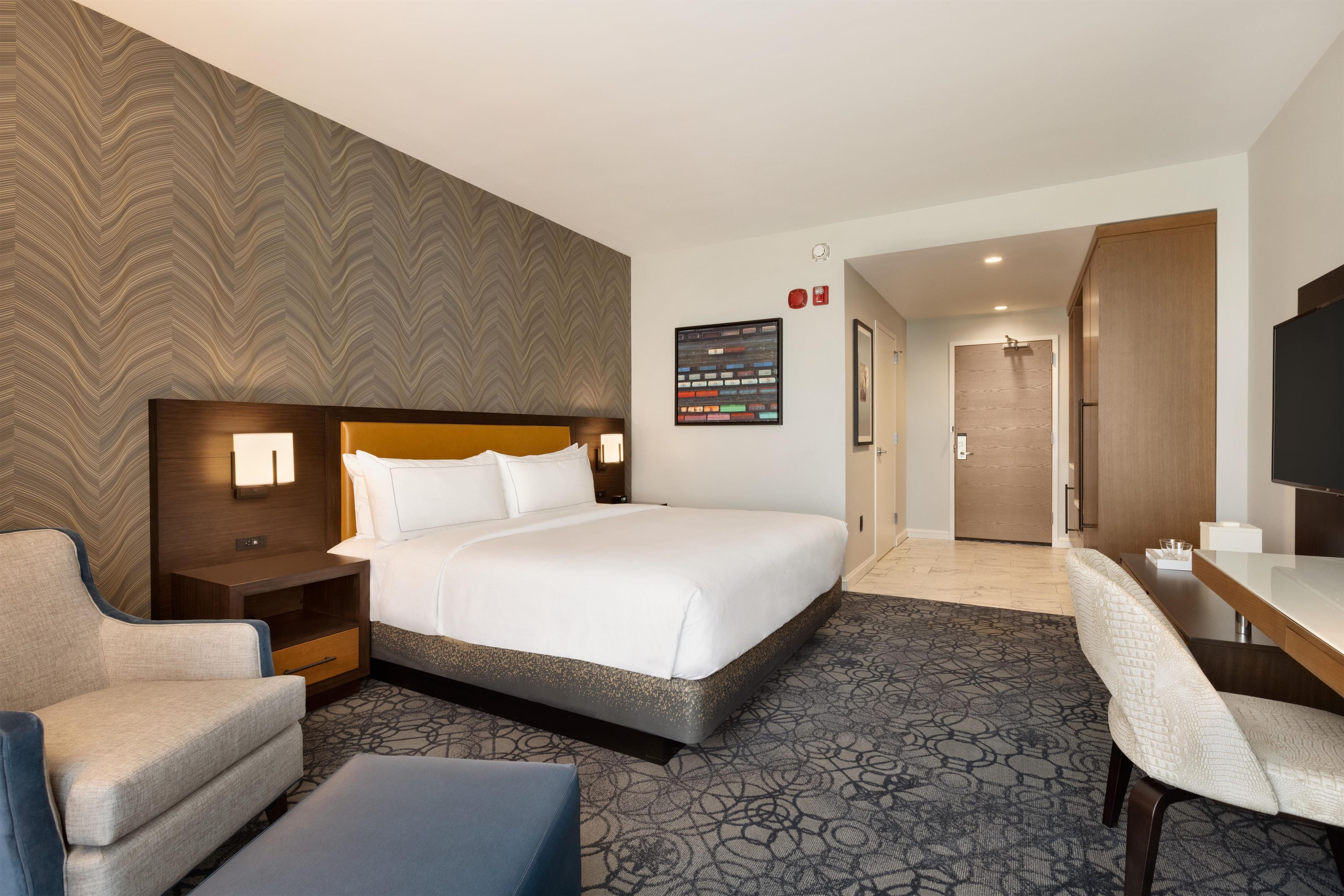 A hotel room with a large bed, a desk, a chair, a nightstand, and a wall-mounted TV.