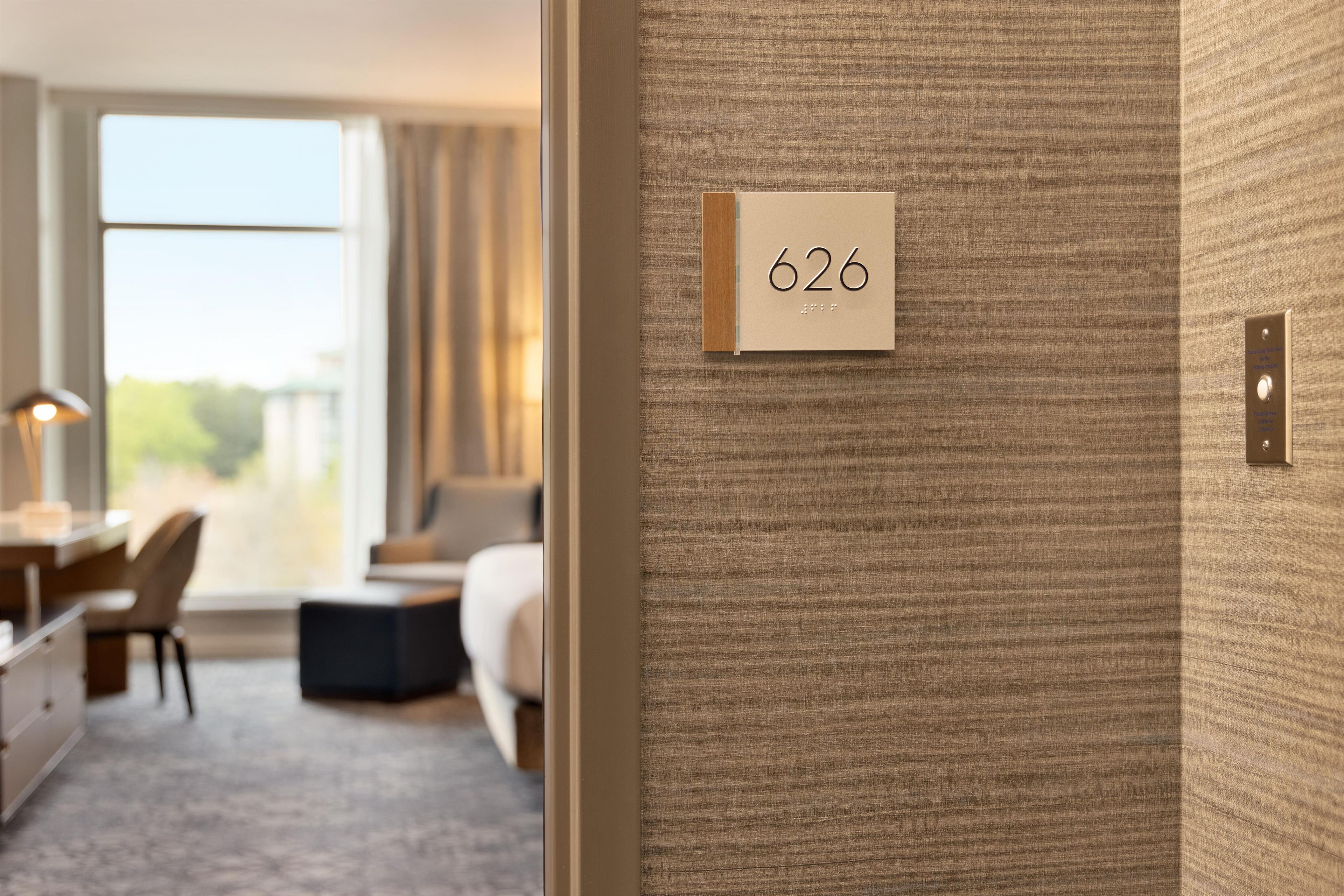 Una habitación de hotel con un ventanal grande, un escritorio y una cama. Un letrero con el número 626 es visible en la pared.
