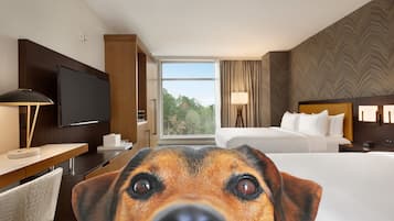 A hotel room with a large bed, a TV, a desk with a lamp, and a view of trees through the window.