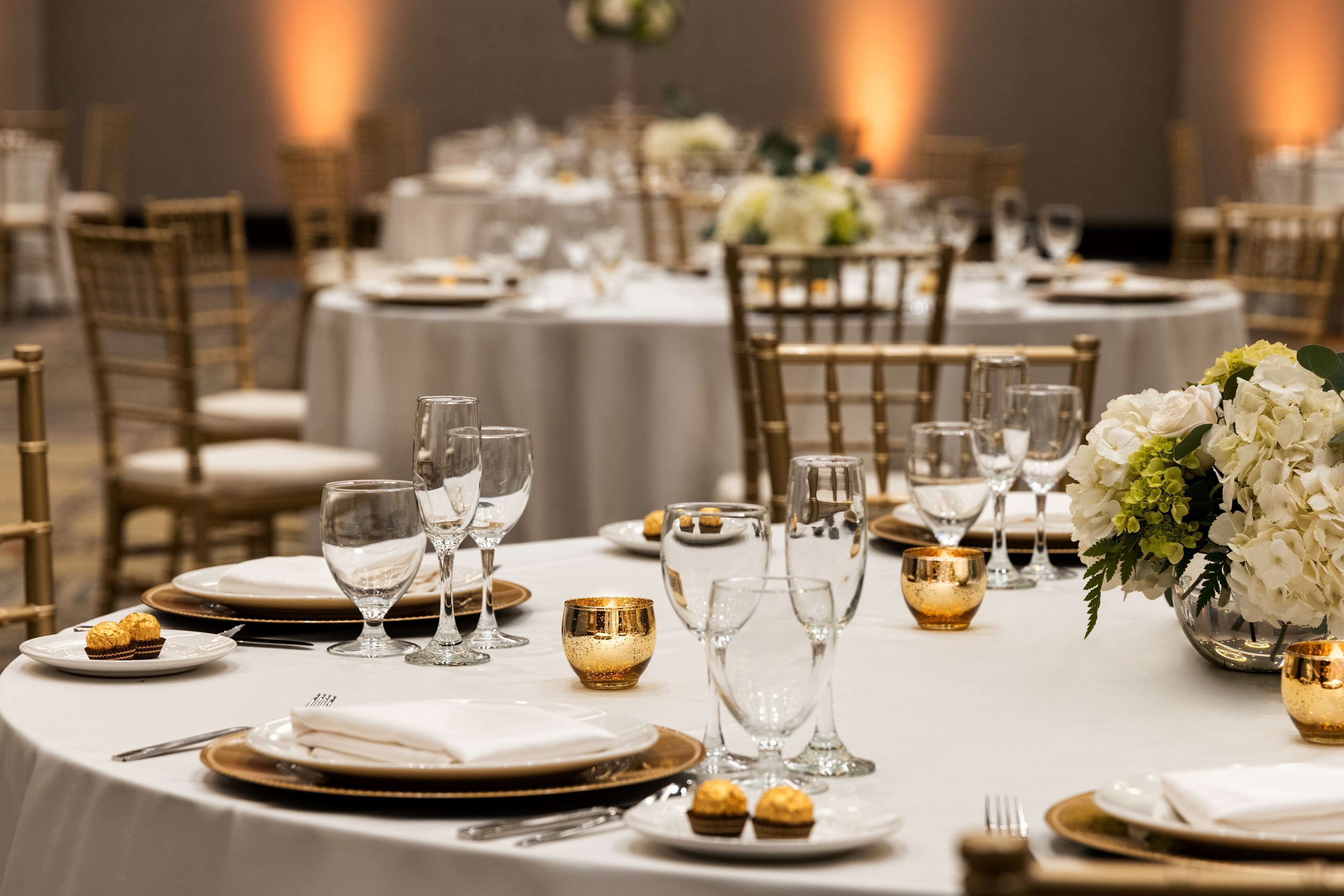 A banquet hall with round tables set for a formal event, featuring gold and white tableware, glassware, and floral centerpieces.
