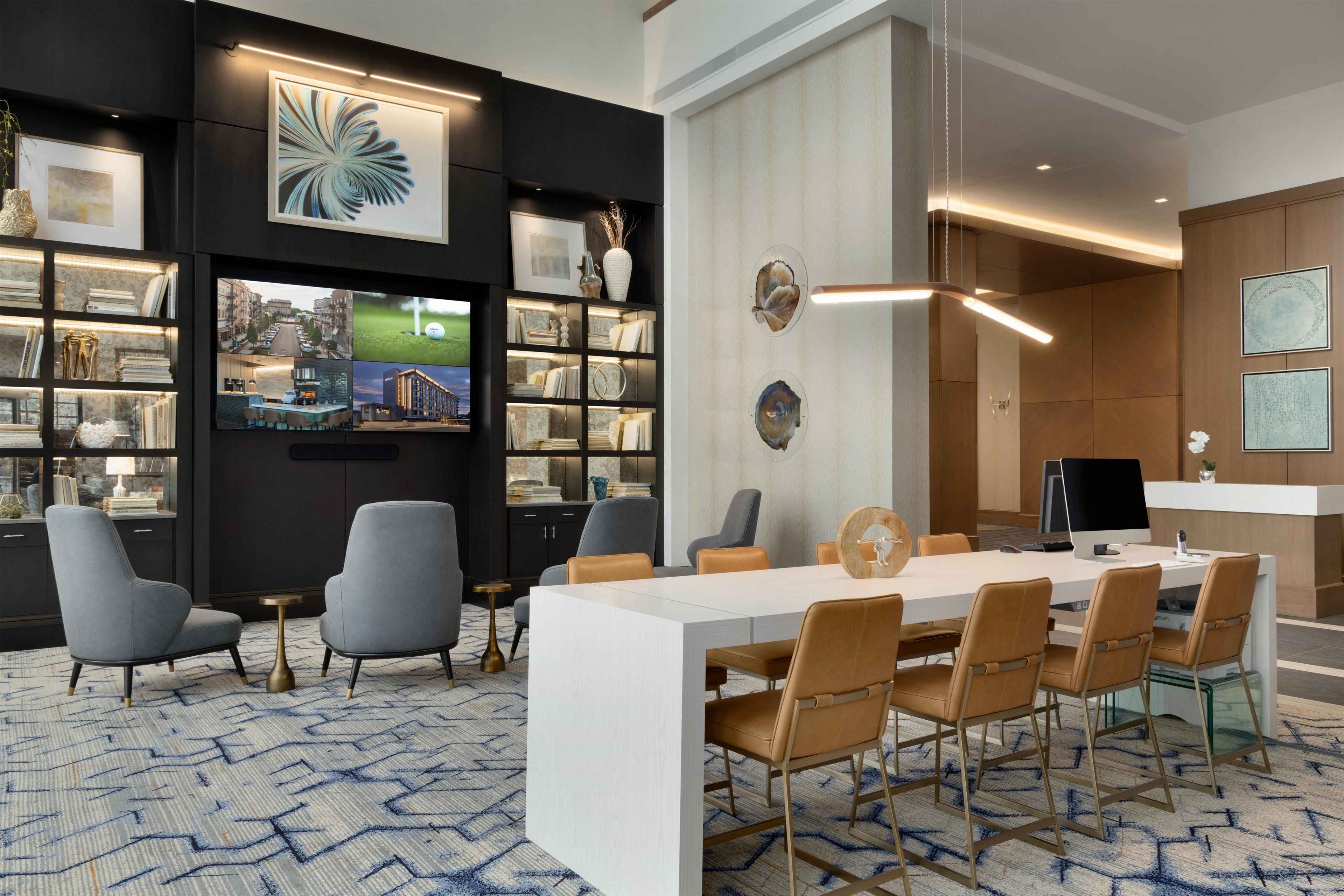 A modern hotel room with a large white desk, multiple chairs, a bookshelf, and a large window.