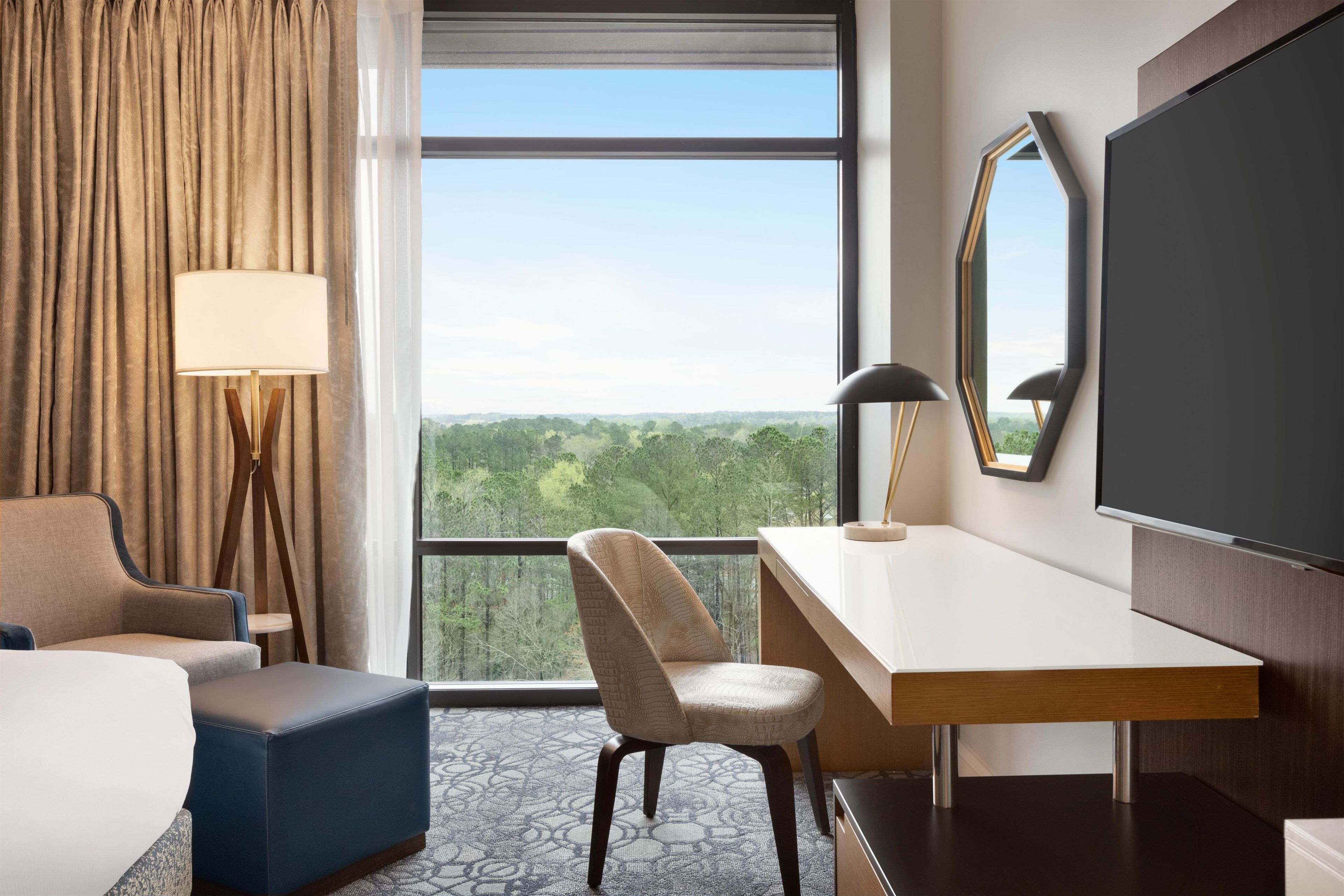 Une chambre d’hôtel avec une grande fenêtre, un bureau, une chaise, un lit et un téléviseur.