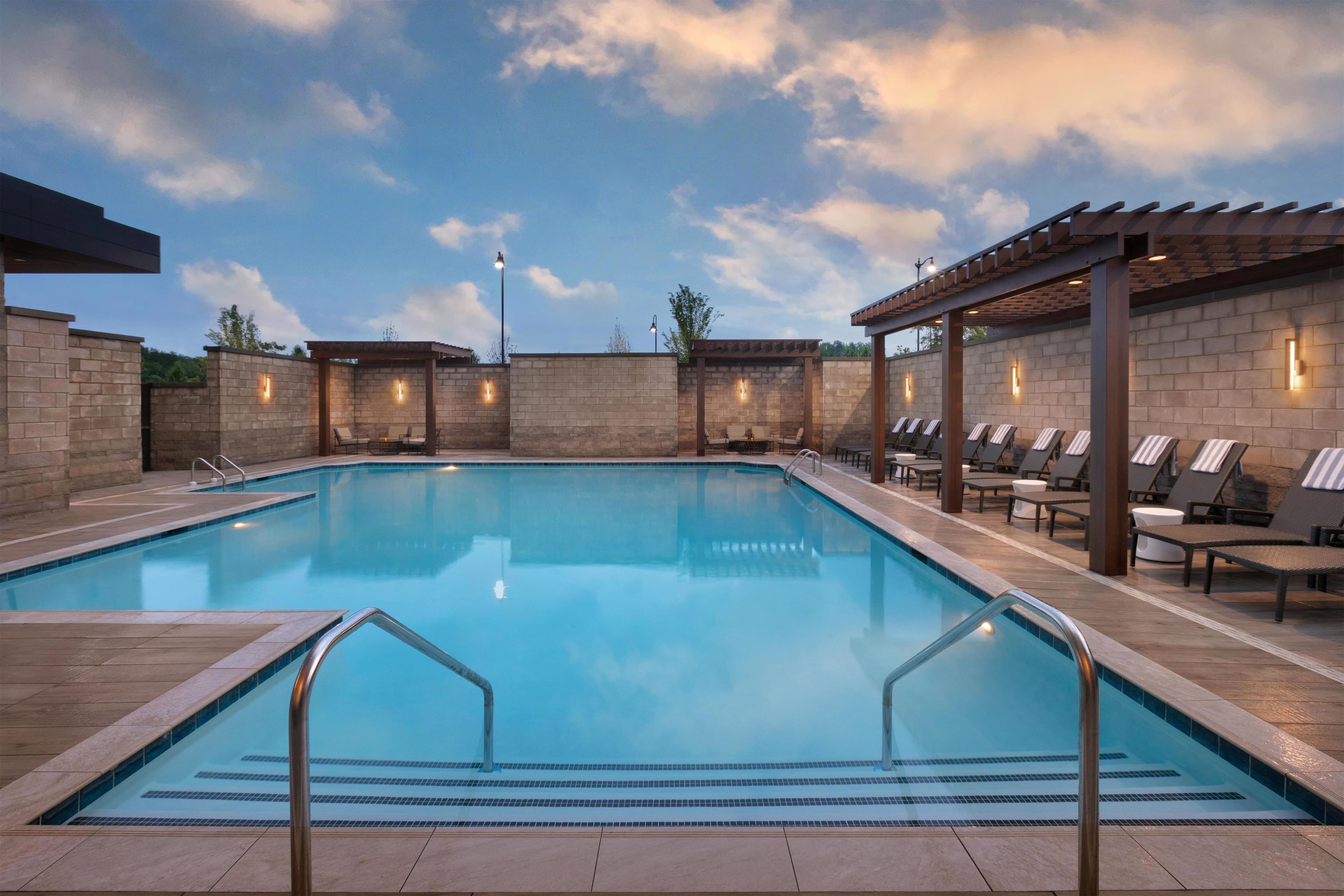 A rectangular swimming pool with a tiled deck, surrounded by lounge chairs and a pergola structure.