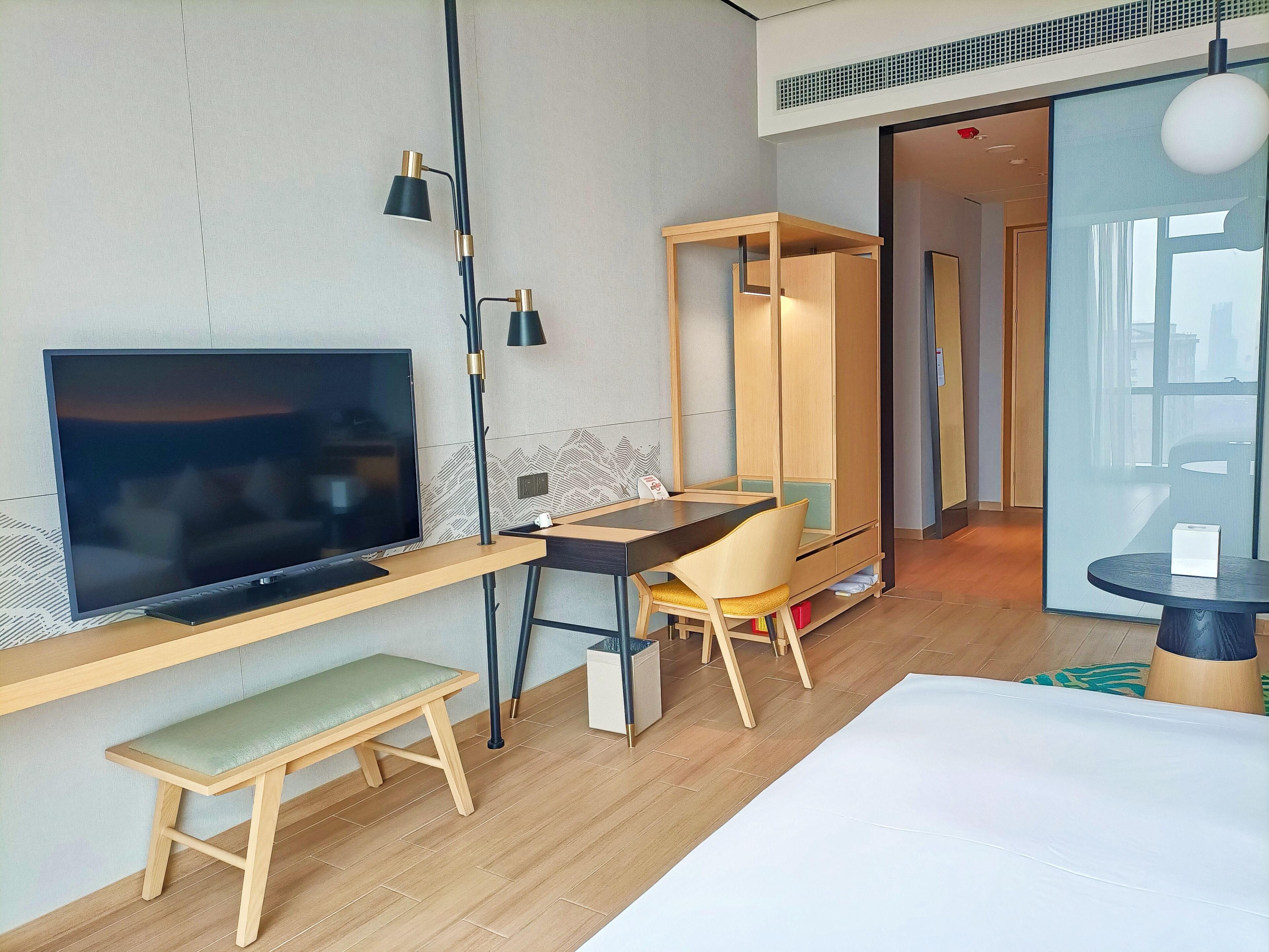 A modern hotel room with a flat-screen TV, a wooden desk, a chair, a bench, and a round table.