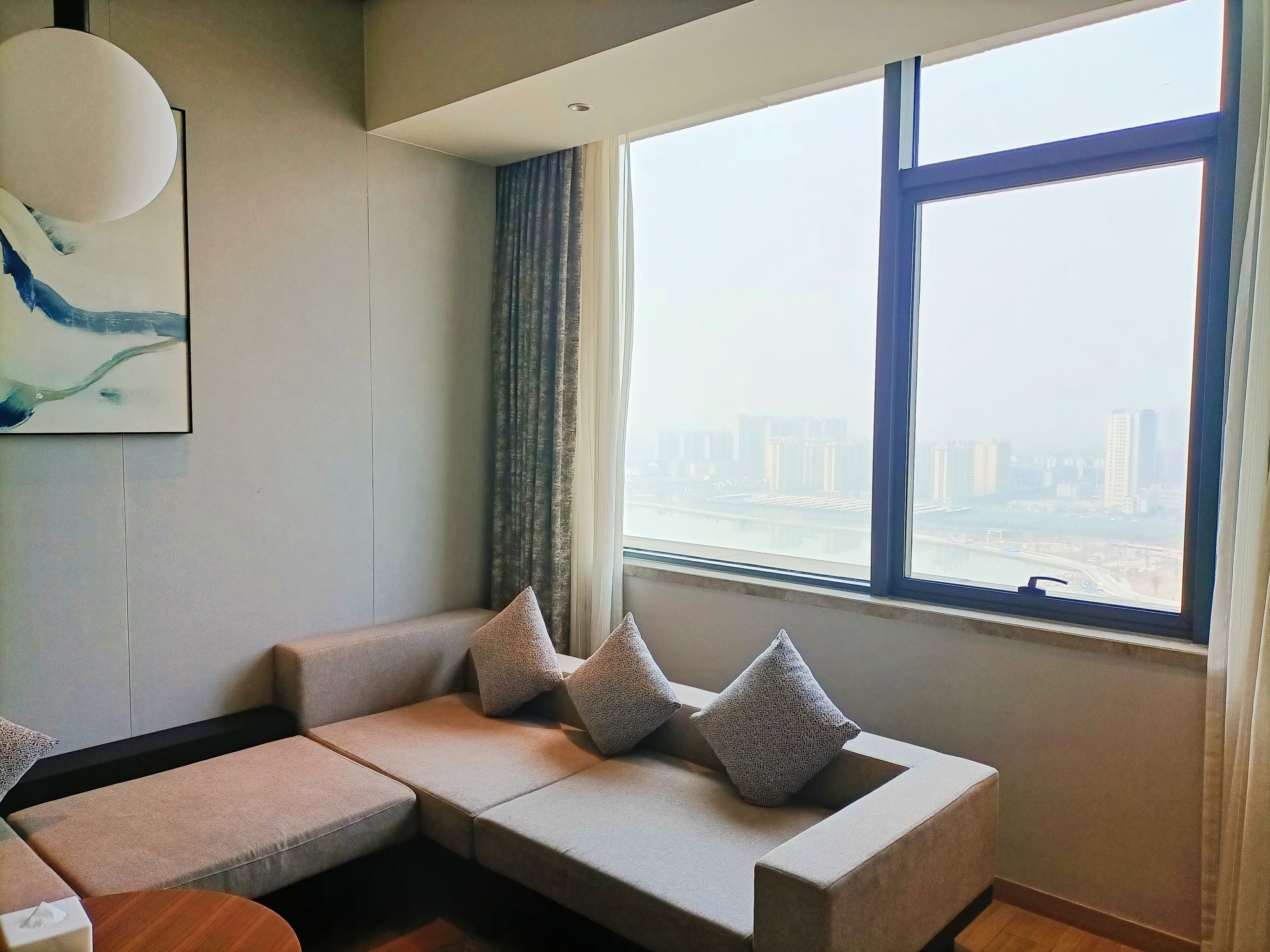 A modern living room with a sofa, pillows, a painting, and a large window with a city view.