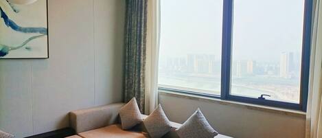 A modern living room with a sofa, pillows, a painting, and a large window with a city view.