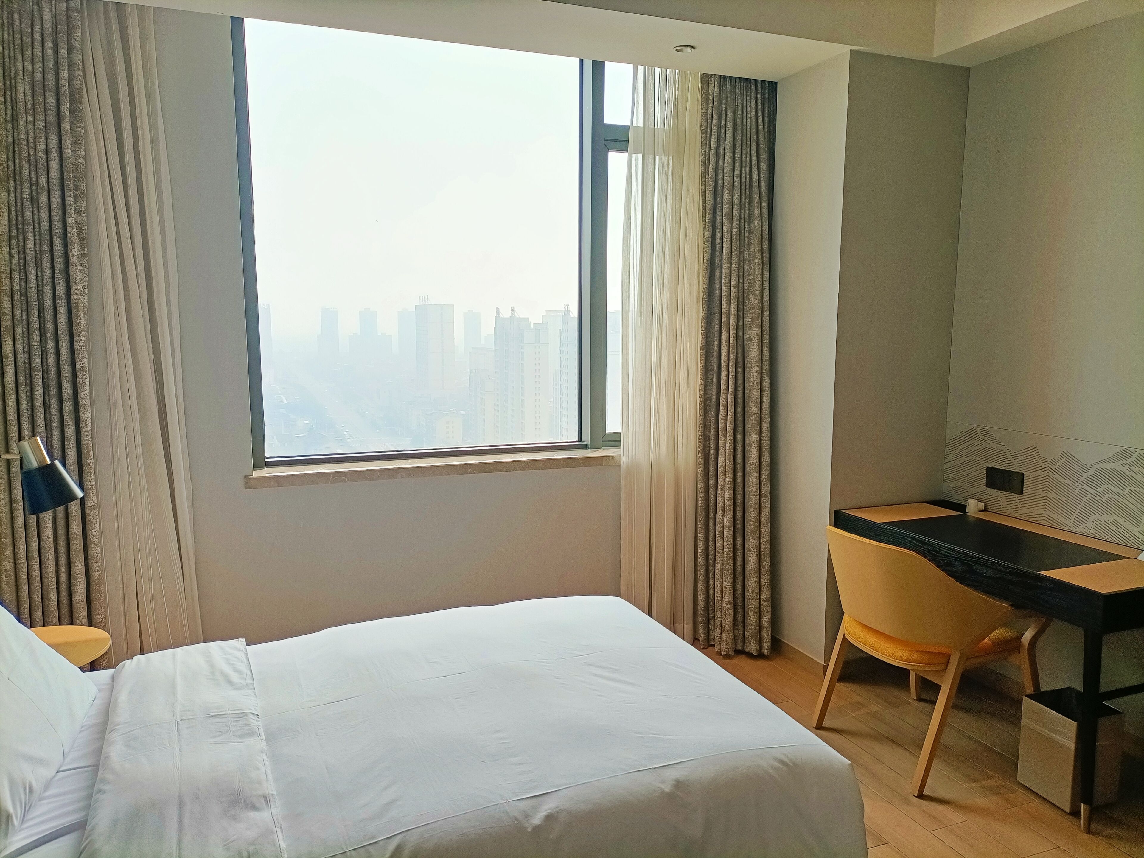 A hotel room with a large window, a bed, a desk, and a chair.