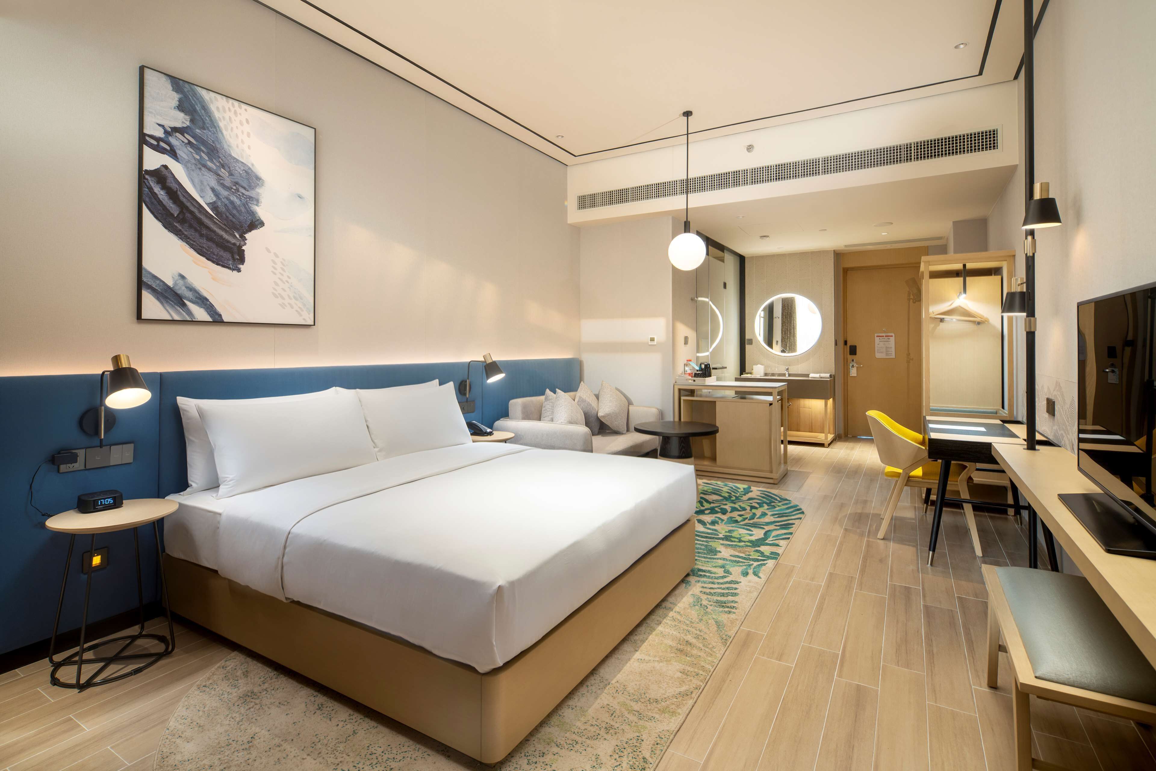 Habitación de hotel con una cama grande, mesita de noche con lámpara, escritorio con silla, espejo y televisión.