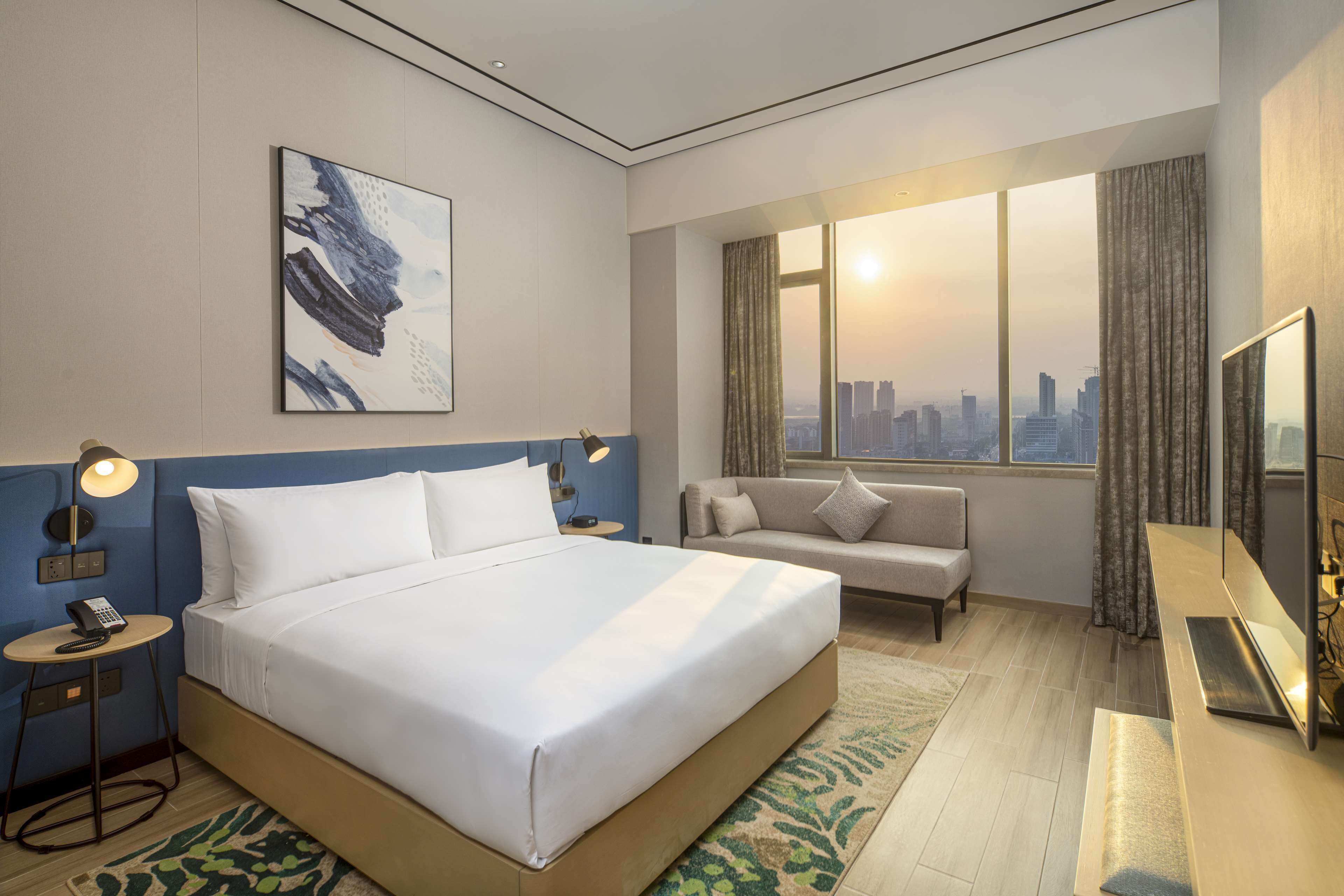 A hotel room with a large bed, a sofa, a desk with a mirror, a city view, and a painting on the wall.