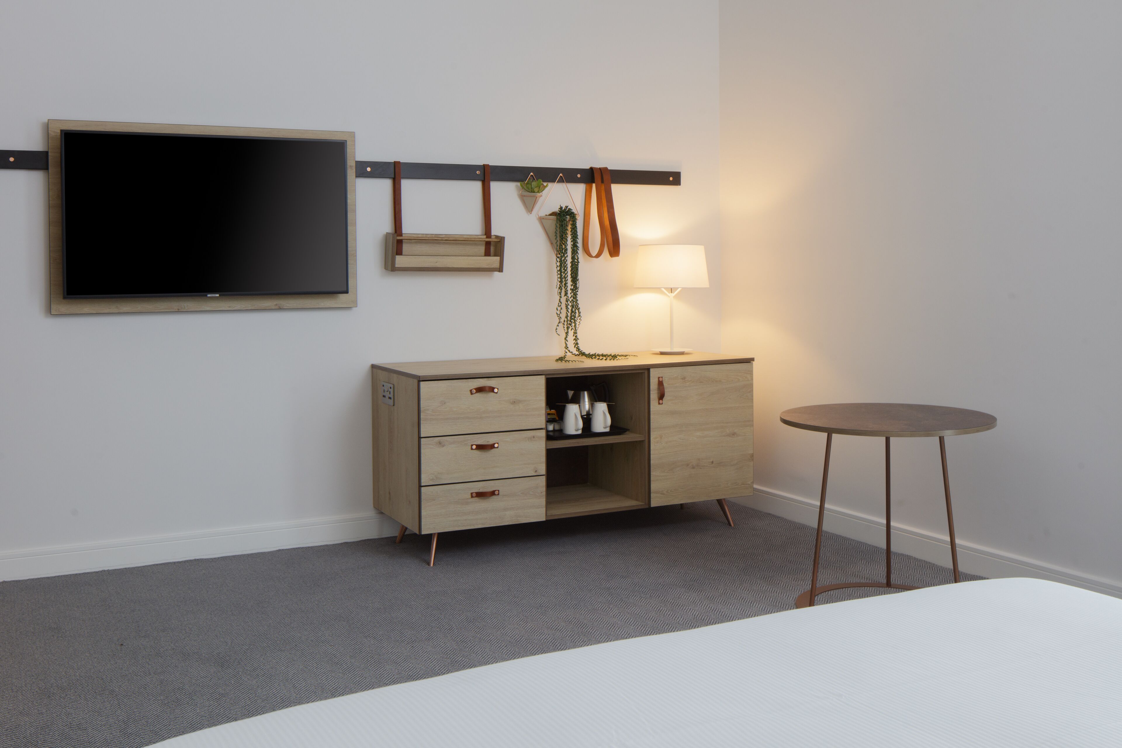 A hotel room with a bed, a wooden dresser with drawers, a flat-screen TV mounted on the wall, a small table, and a lamp.
