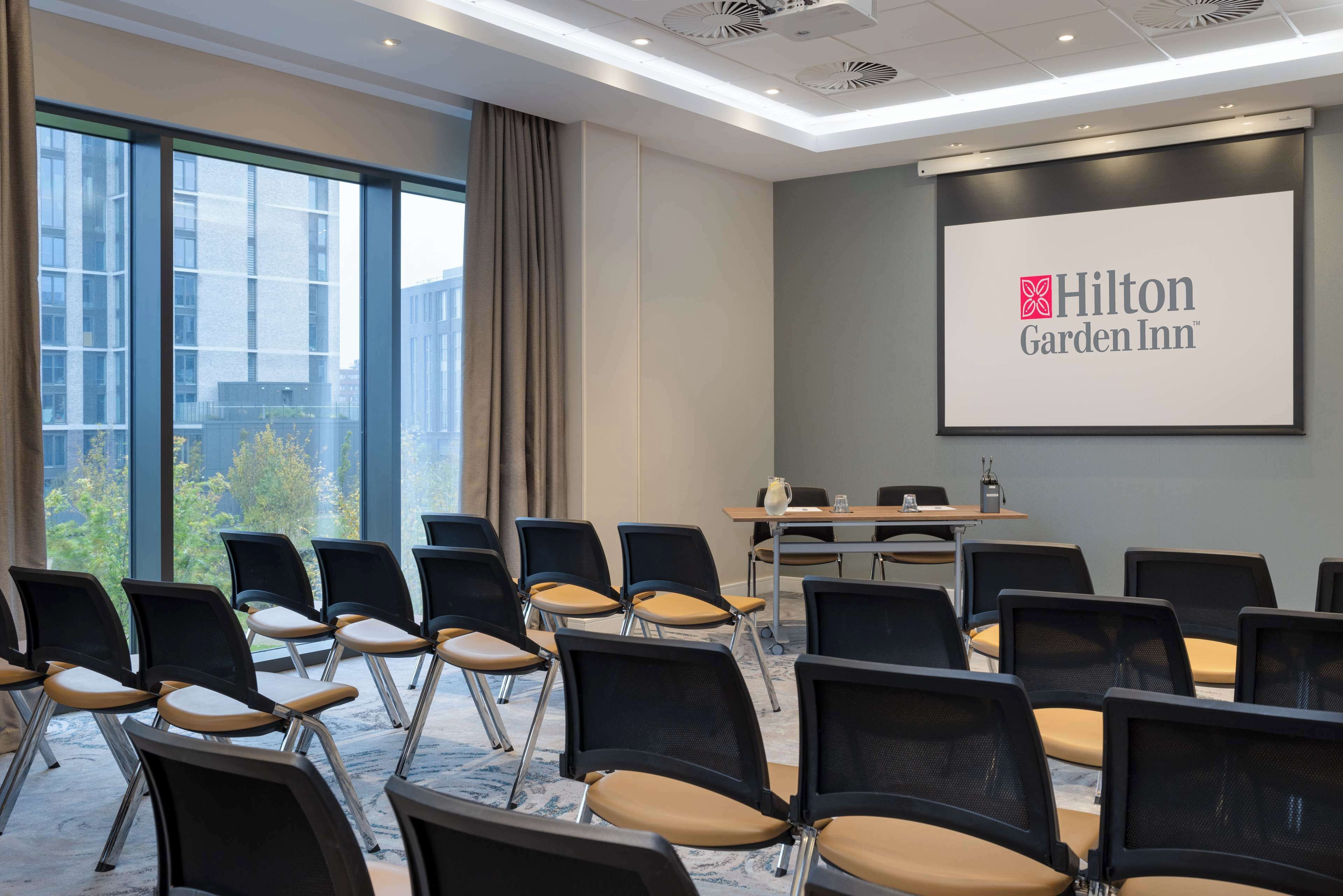 Een vergaderzaal met rijen stoelen, een projectorscherm waarop het Hilton Garden Inn logo staat, en een groot raam met uitzicht op gebouwen en bomen.