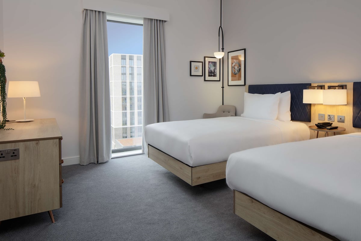 Een hotelkamer met twee bedden, een bureau, een lamp, een nachtkastje en een raam met gordijnen.