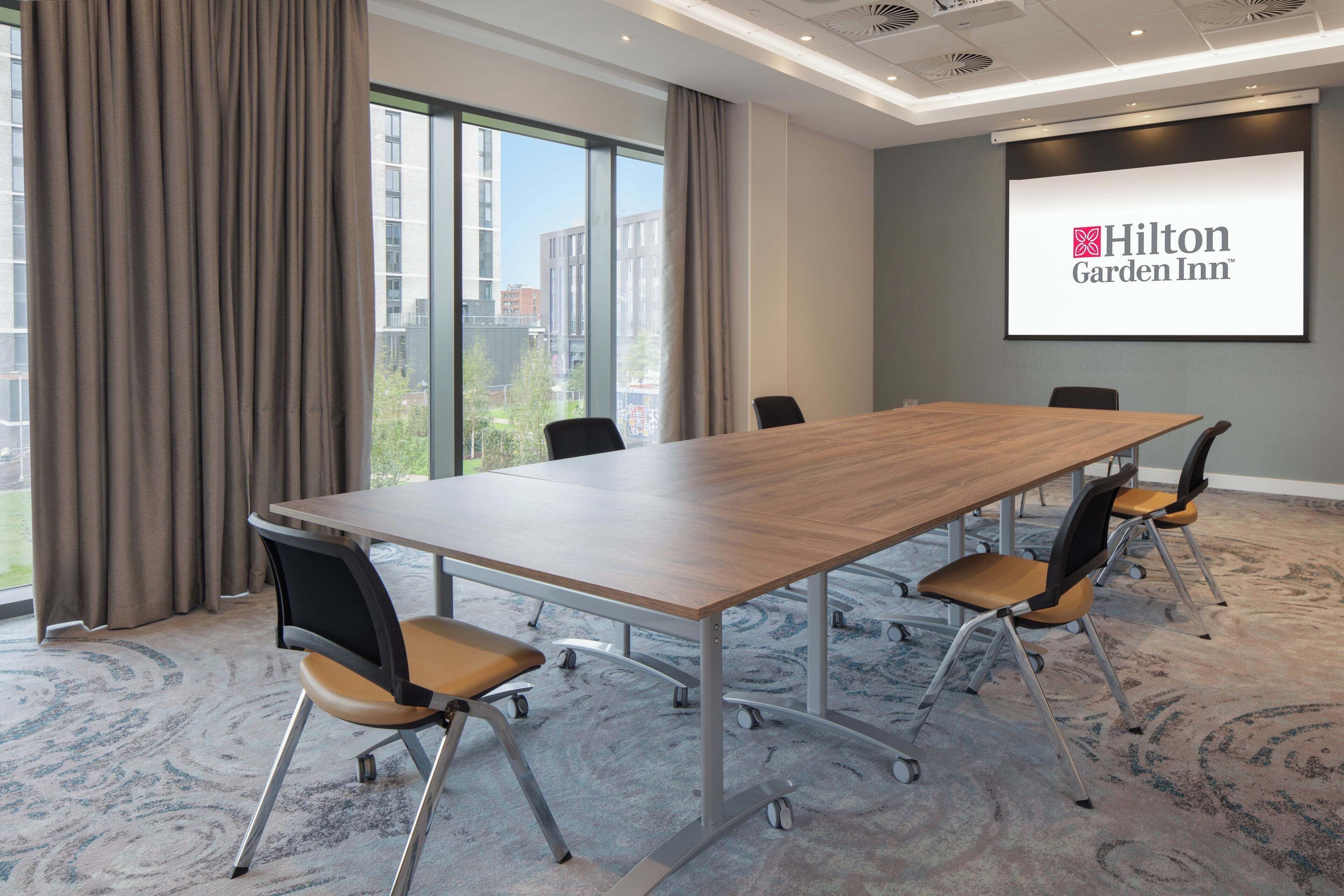 A conference room with a long table, chairs, a projector screen displaying the Hilton Garden Inn logo, and large windows with curtains.