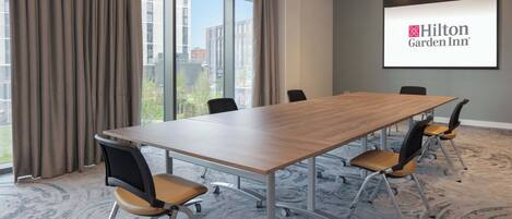 A conference room with a long table, chairs, a projector screen displaying the Hilton Garden Inn logo, and large windows with curtains.