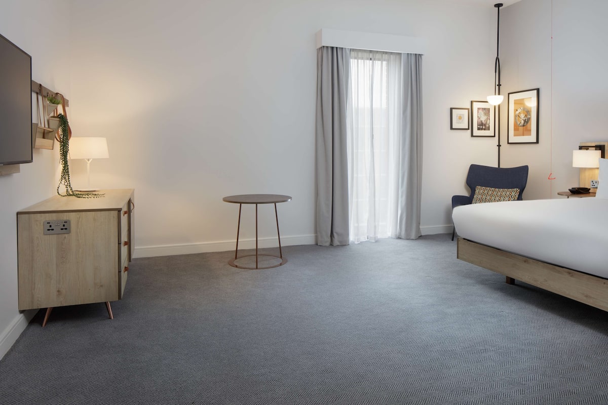 Een hotelkamer met een bed, een nachtkastje met lamp, een klein rond tafeltje, een aan de muur bevestigde televisie en een raam met gordijnen.