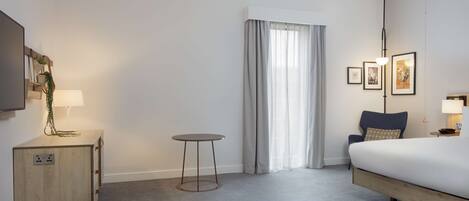 A hotel room with a bed, a bedside table with a lamp, a small round table, a wall-mounted TV, and a window with curtains.