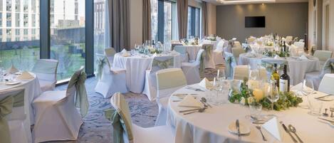 Un salón de banquetes con mesas redondas dispuestas para comer, amplias ventanas y un televisor de pantalla plana en la pared.
