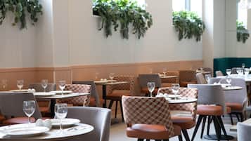 Un restaurante con mesas redondas y sillas a cuadros, con plantas verdes en las paredes y una zona con asientos tipo cabina color verde azulado.