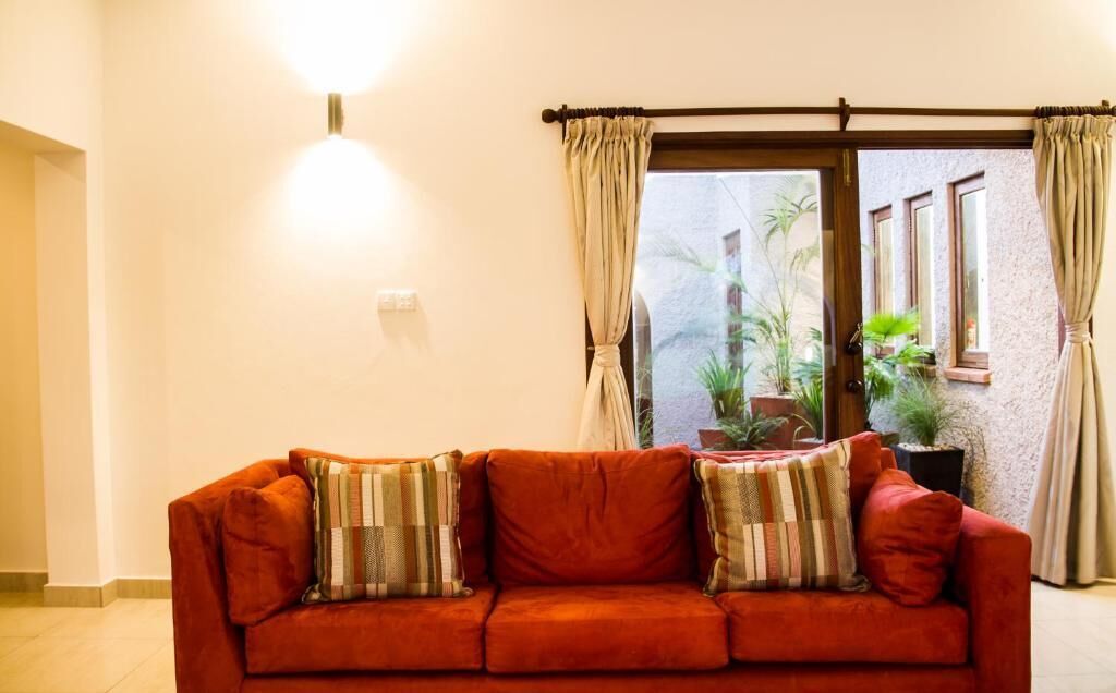 Ein rotes Sofa mit gestreiften Kissen in einem Raum mit großem Fenster, Vorhängen und einer Wandleuchte.
