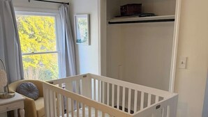 Et børneværelse med tremmeseng, skiftemøbel, skab og et vindue med gardiner.