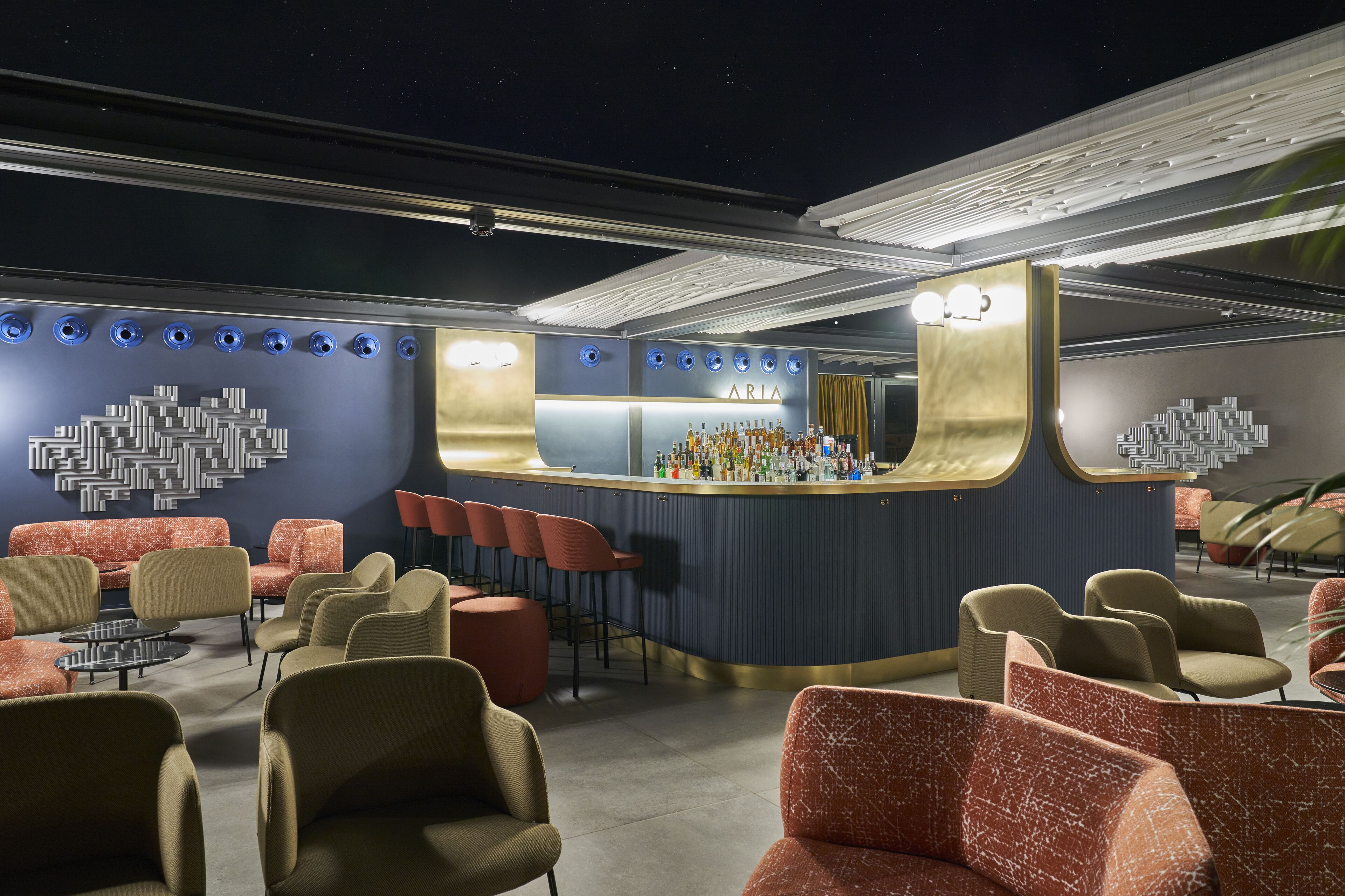 A bar area with a curved counter, seating with red and beige chairs, and a wall with a geometric light fixture.