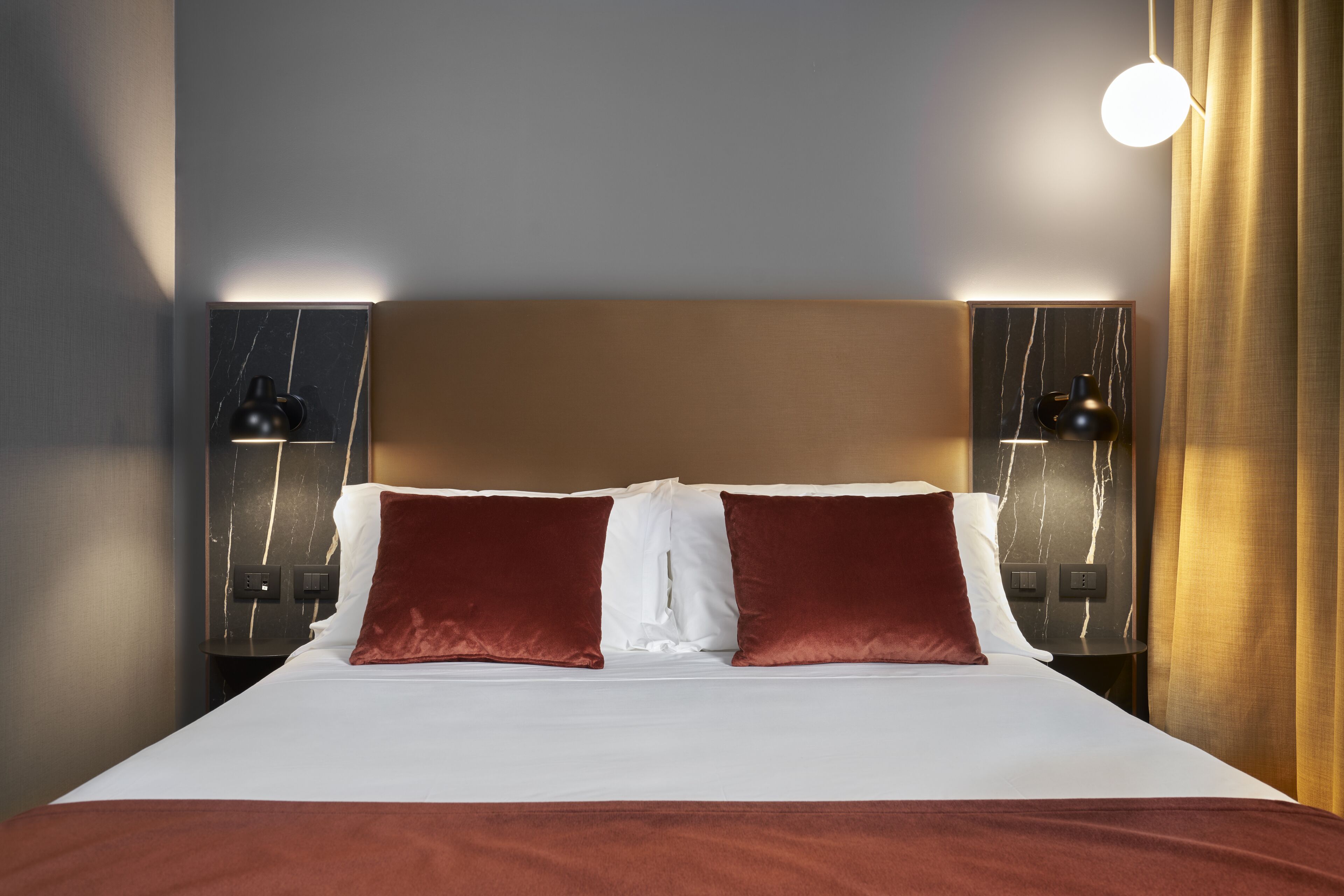 Habitación de hotel con una cama grande, dos lámparas de noche, un cabecero de mármol y una cortina a la derecha.