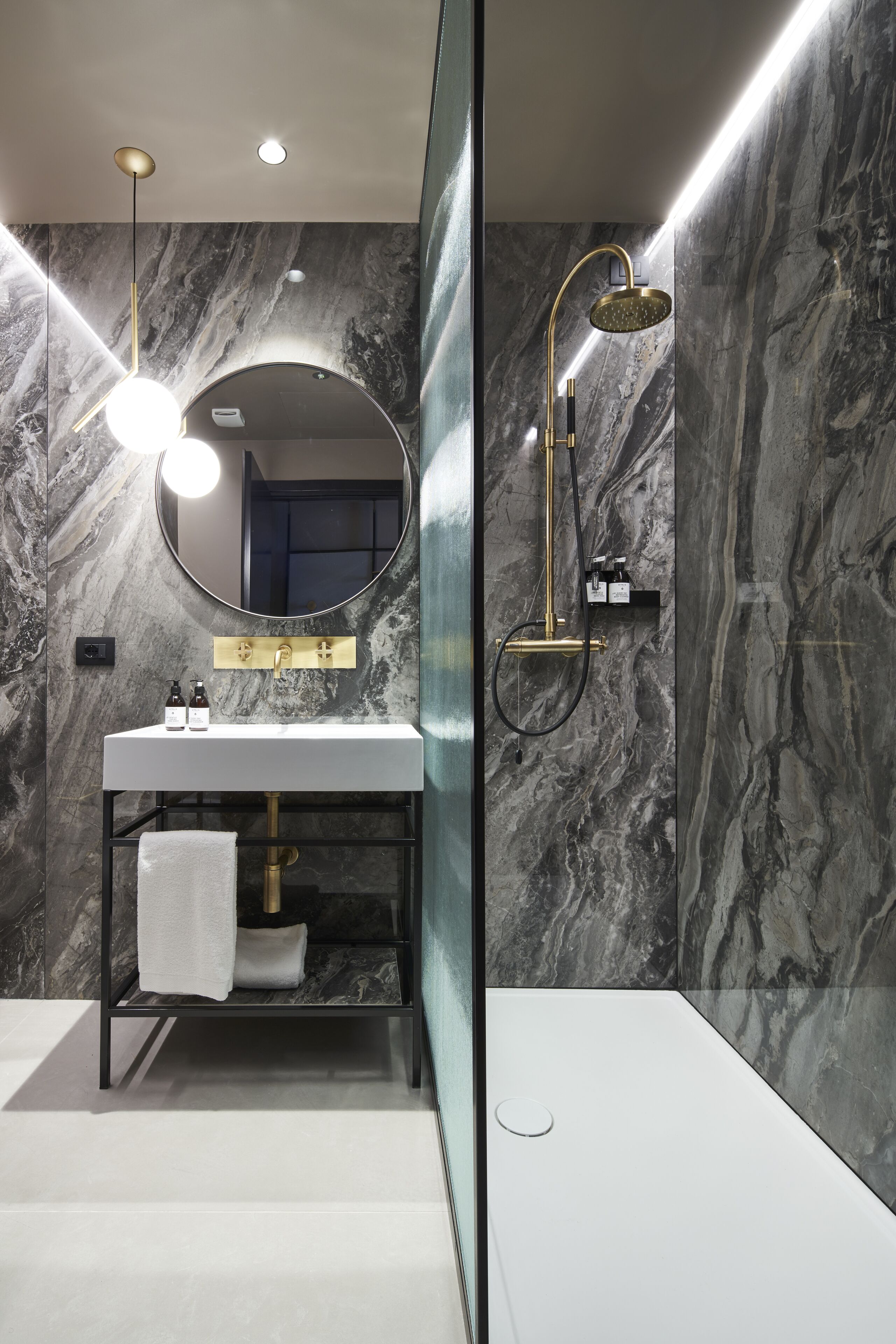 A modern bathroom with a marble wall, a round mirror, a white sink, and a gold showerhead.