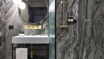 A modern bathroom with a marble wall, a round mirror, a white sink, and a gold showerhead.