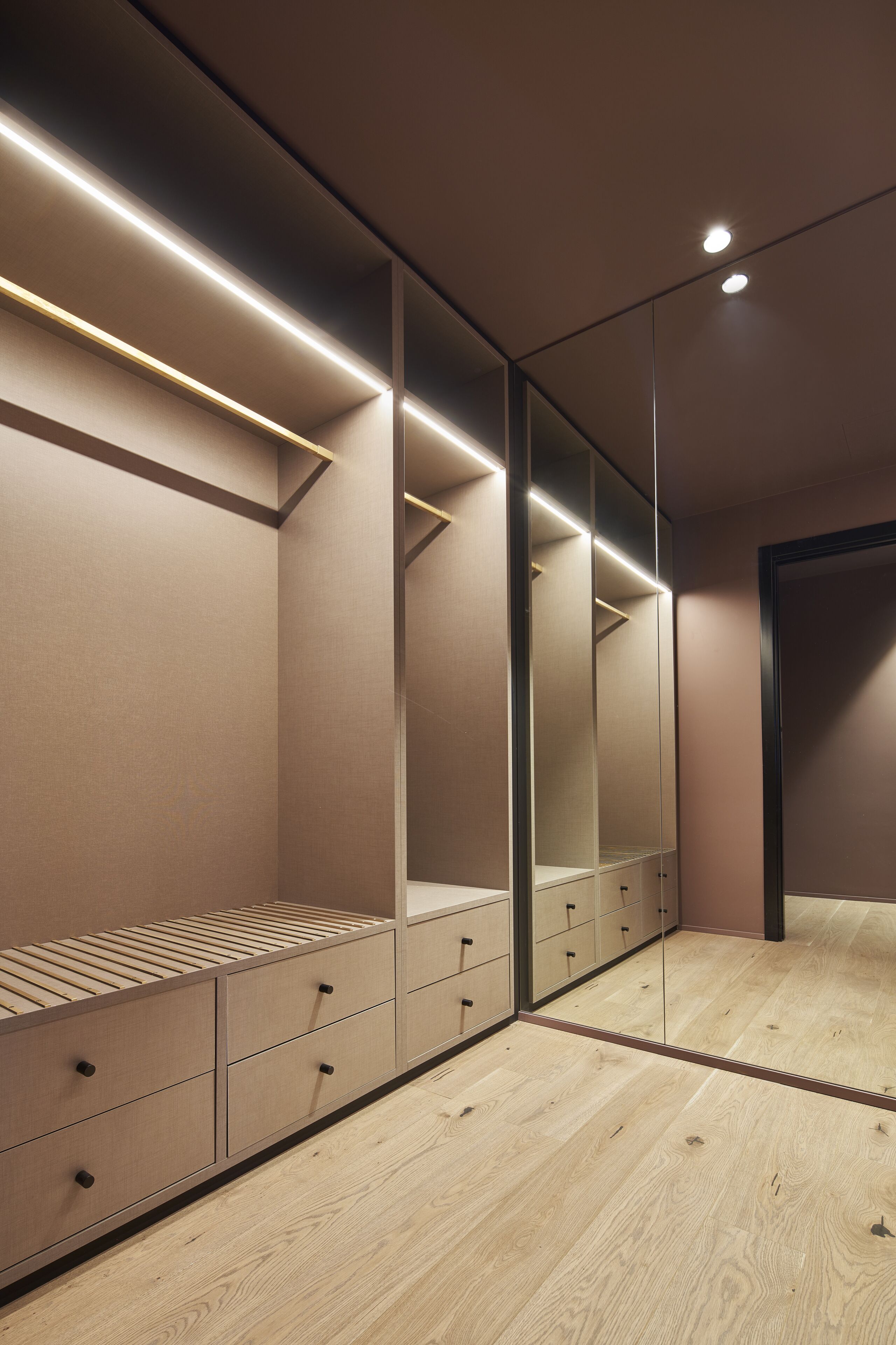 A modern bedroom with a wooden floor, built-in wardrobe with drawers and shelves, and a large mirror.