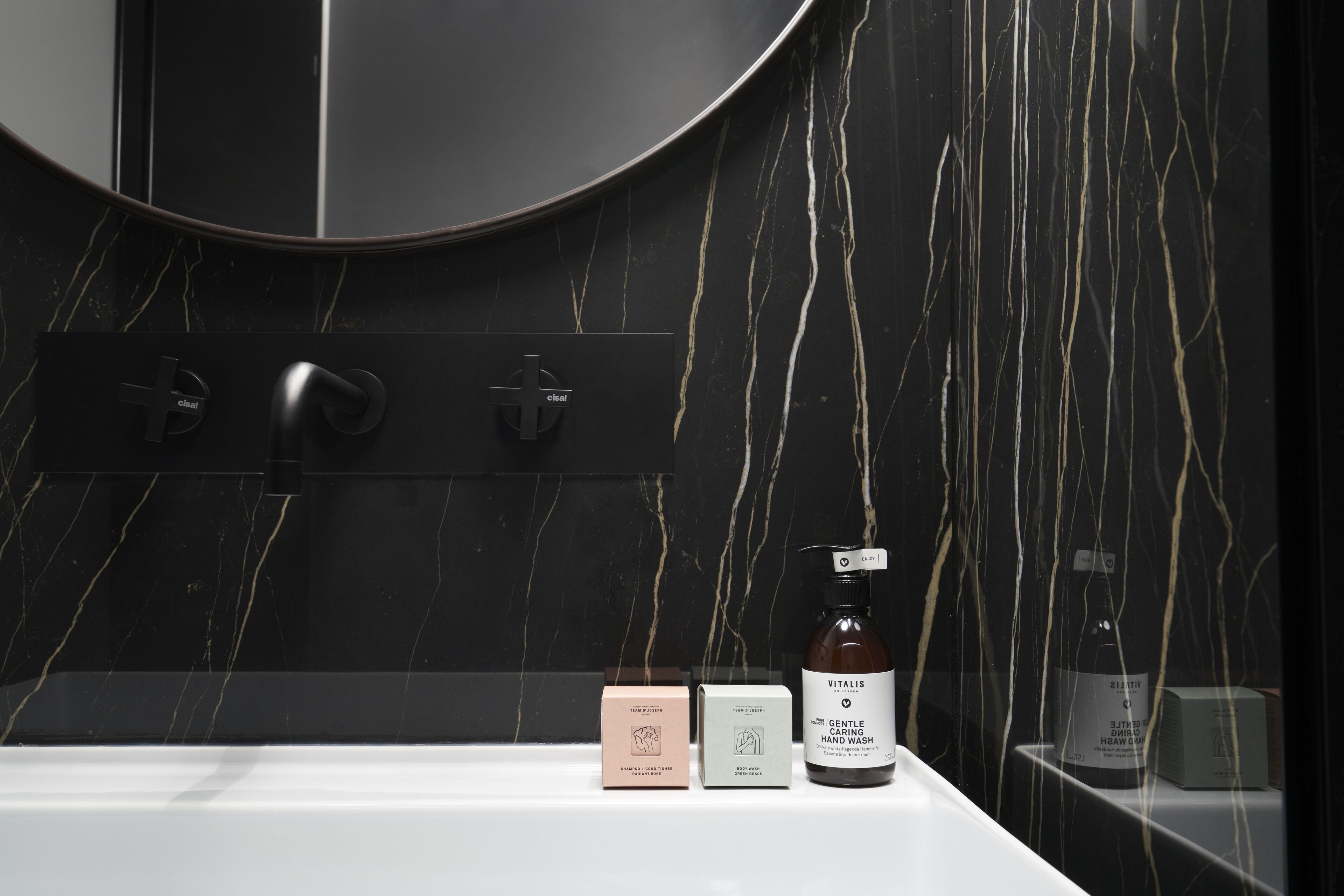 Un baño con pared de mármol negro, un espejo redondo y un lavabo blanco. Hay dos cajas y una botella de jabón de manos sobre el mostrador.